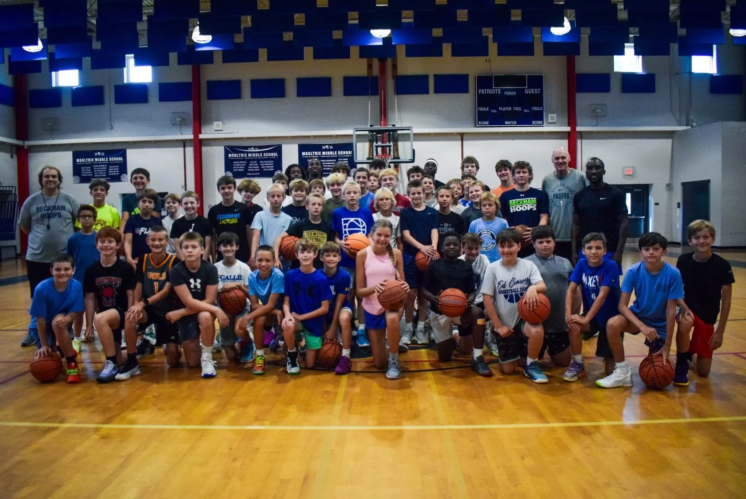 A large group of children and a few adults pose for a group photo on a basketball court inside a gymnasium. The children are wearing sports attire, some holding basketballs, and the group is smiling at the camera.