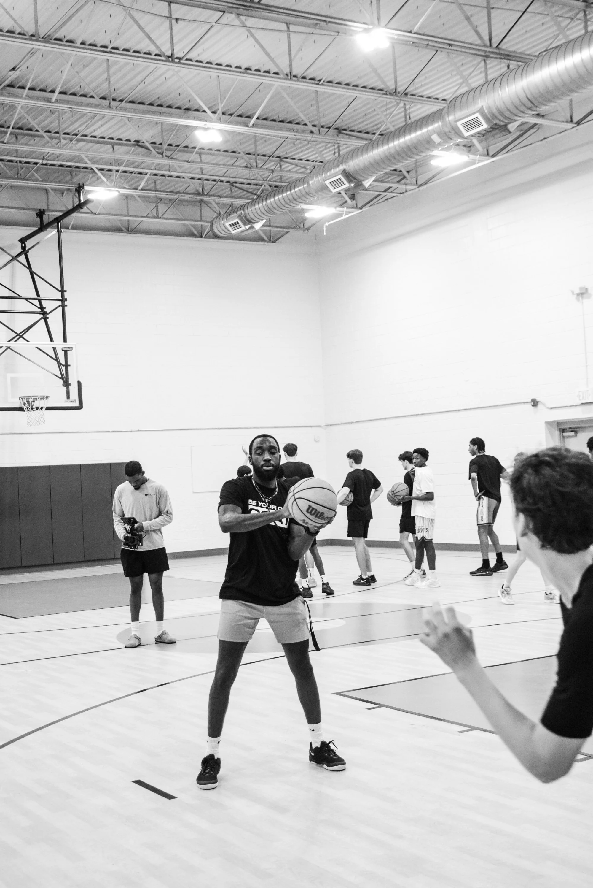 A group of young men practicing basketball in an indoor gymnasium, with one player in the foreground holding a basketball, preparing to shoot or pass.