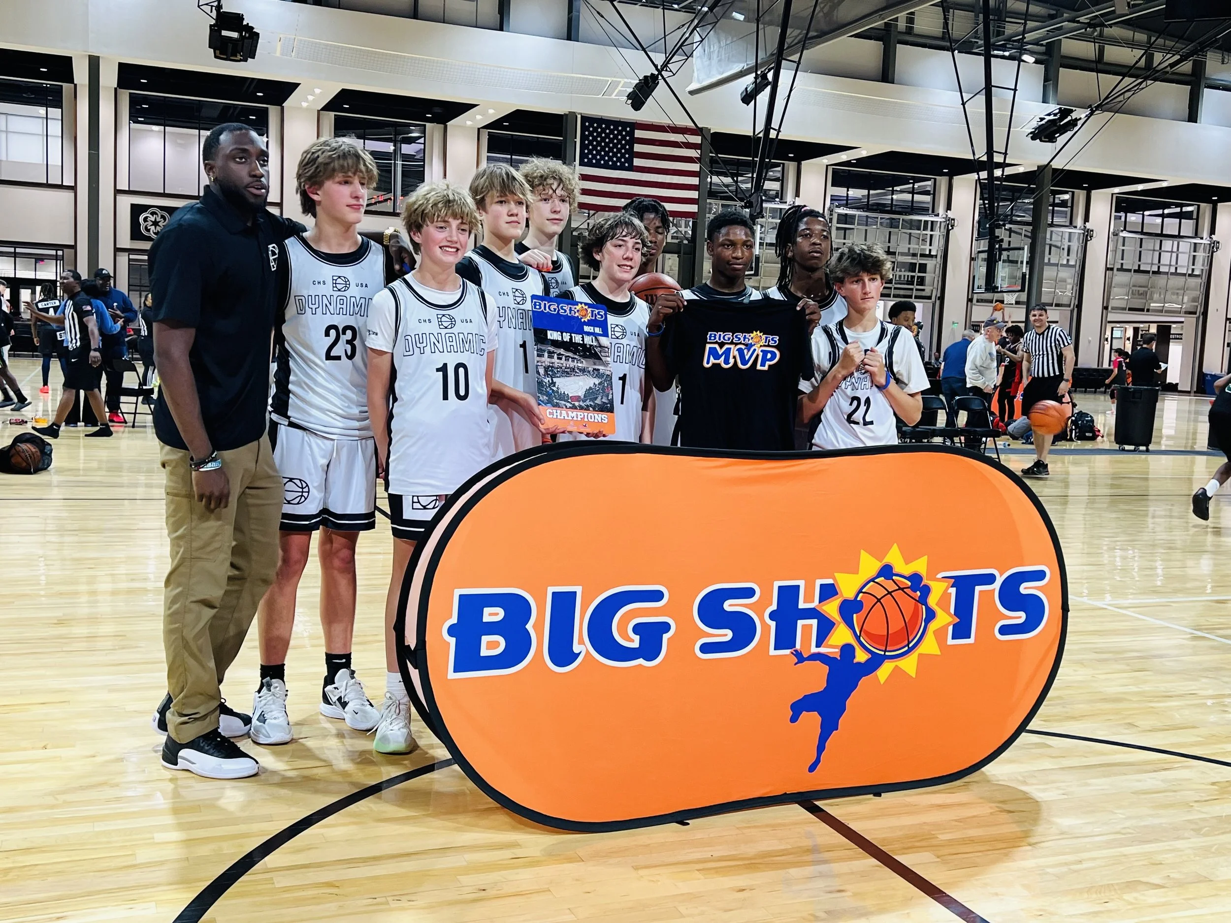 Group of young basketball players and coach posing behind a large orange 'Big Shots' sign at an indoor basketball court. The players are wearing white uniforms with black accents and one player is holding an MVP award. There are other people and play