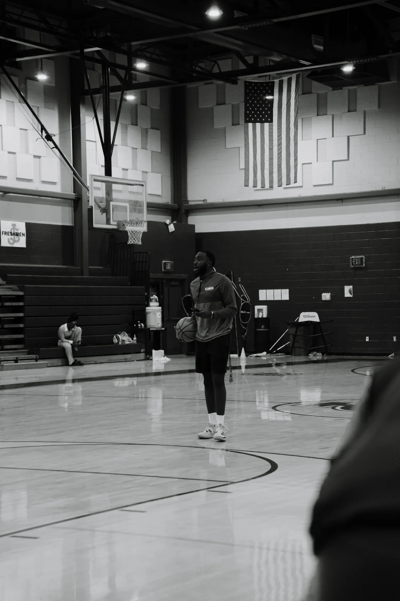 A basketball coach or player standing on an indoor basketball court holding a basketball, with another person sitting on the bleachers in the background. The American flag hangs on the wall behind them.
