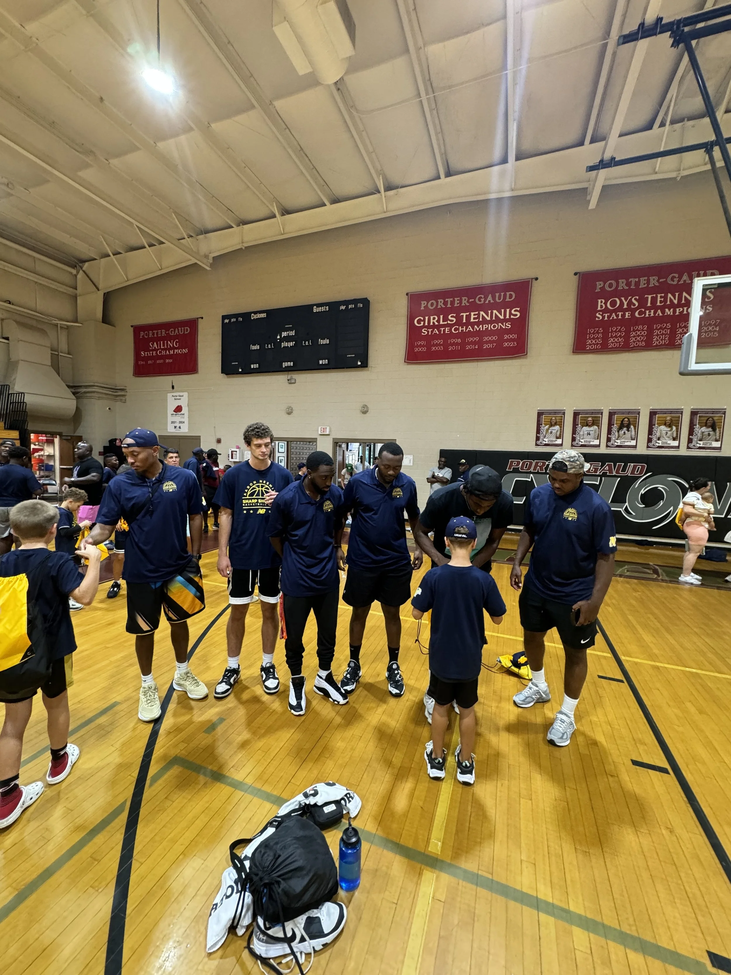A group of children and teenagers in navy blue athletic shirts and shorts standing on a gymnasium floor, with some holding yellow bags, while a few adults in matching shirts are interacting with them. The gym has wooden flooring, sports banners on th