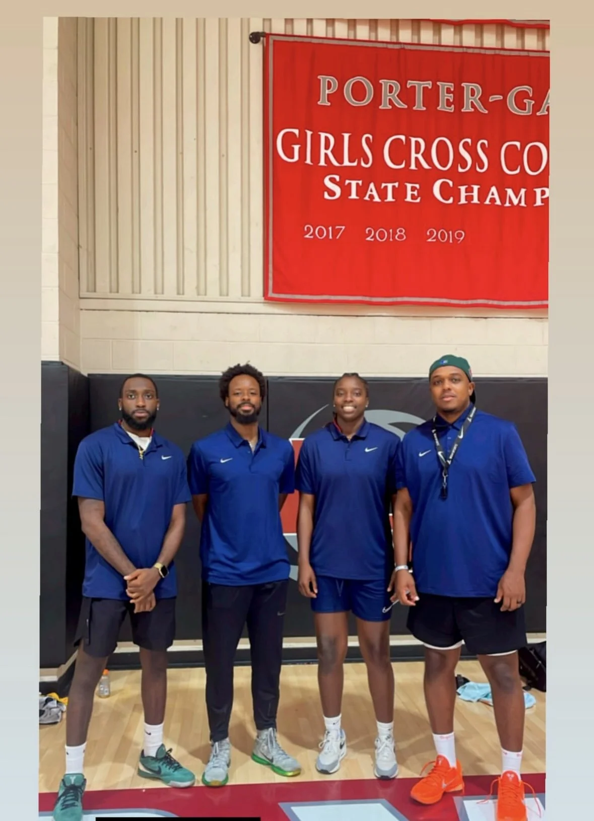 Four athletes standing in front of a red banner that reads 'Porter-Girls Cross Country State Champs 2017, 2018, 2019' inside a gymnasium. The athletes are wearing blue Nike shirts and athletic shorts or pants.