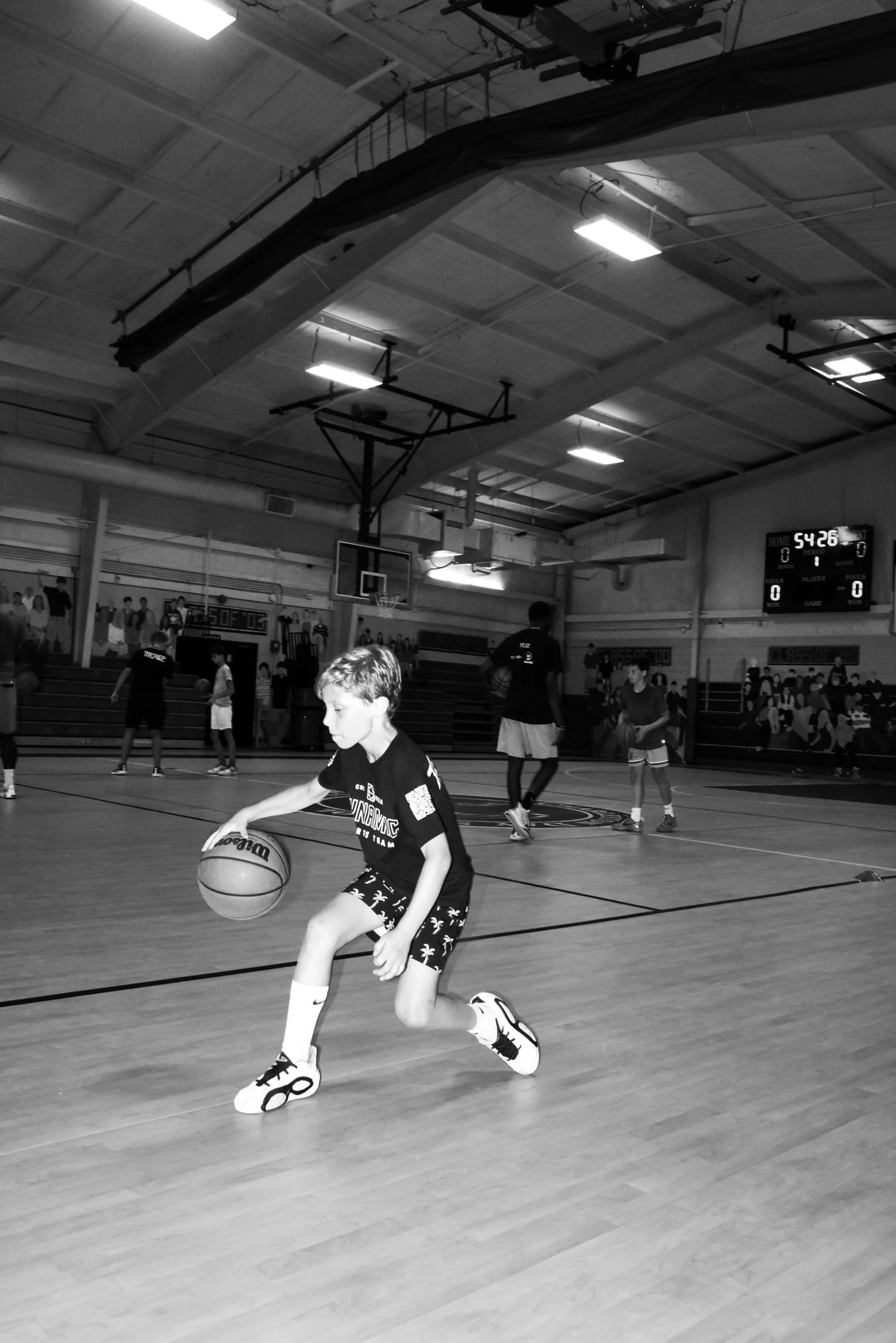 A young boy dribbling a basketball on an indoor court during a youth basketball practice or game, with other children and spectators in the background.