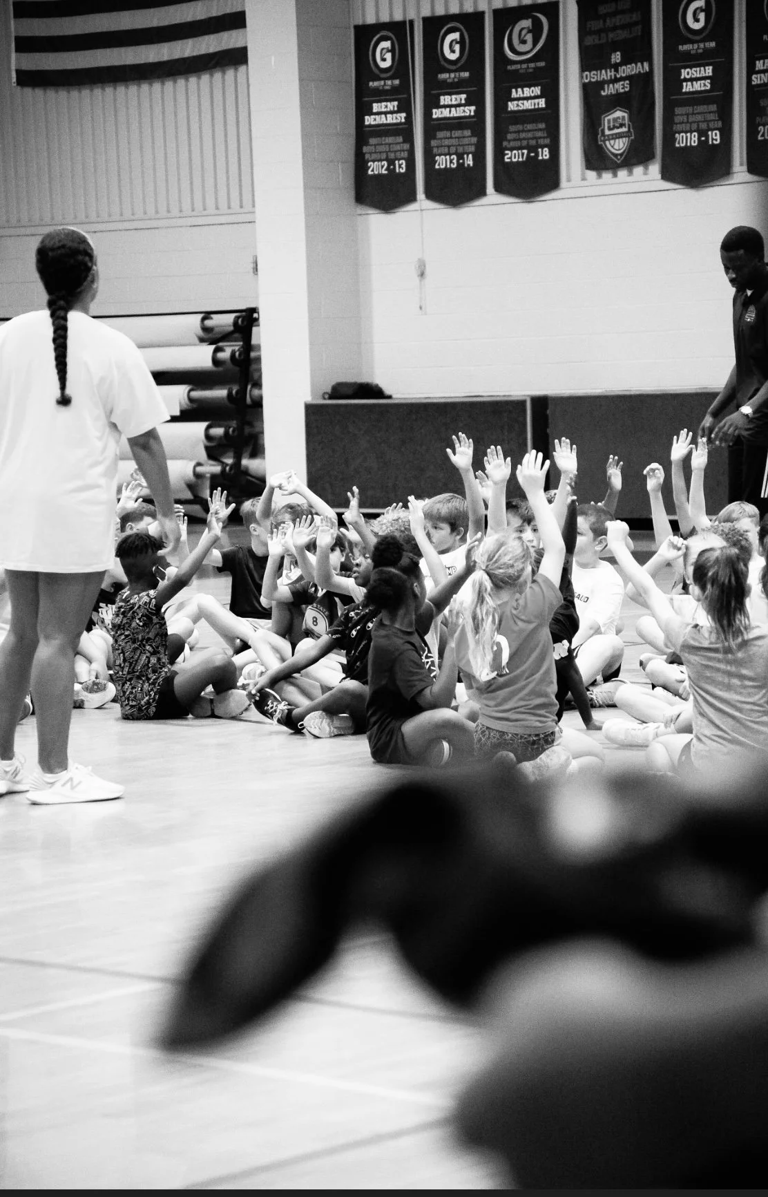 A group of children sitting cross-legged on the floor of a gymnasium, raising their hands, with two instructors standing nearby, and banners hanging on the wall in the background.