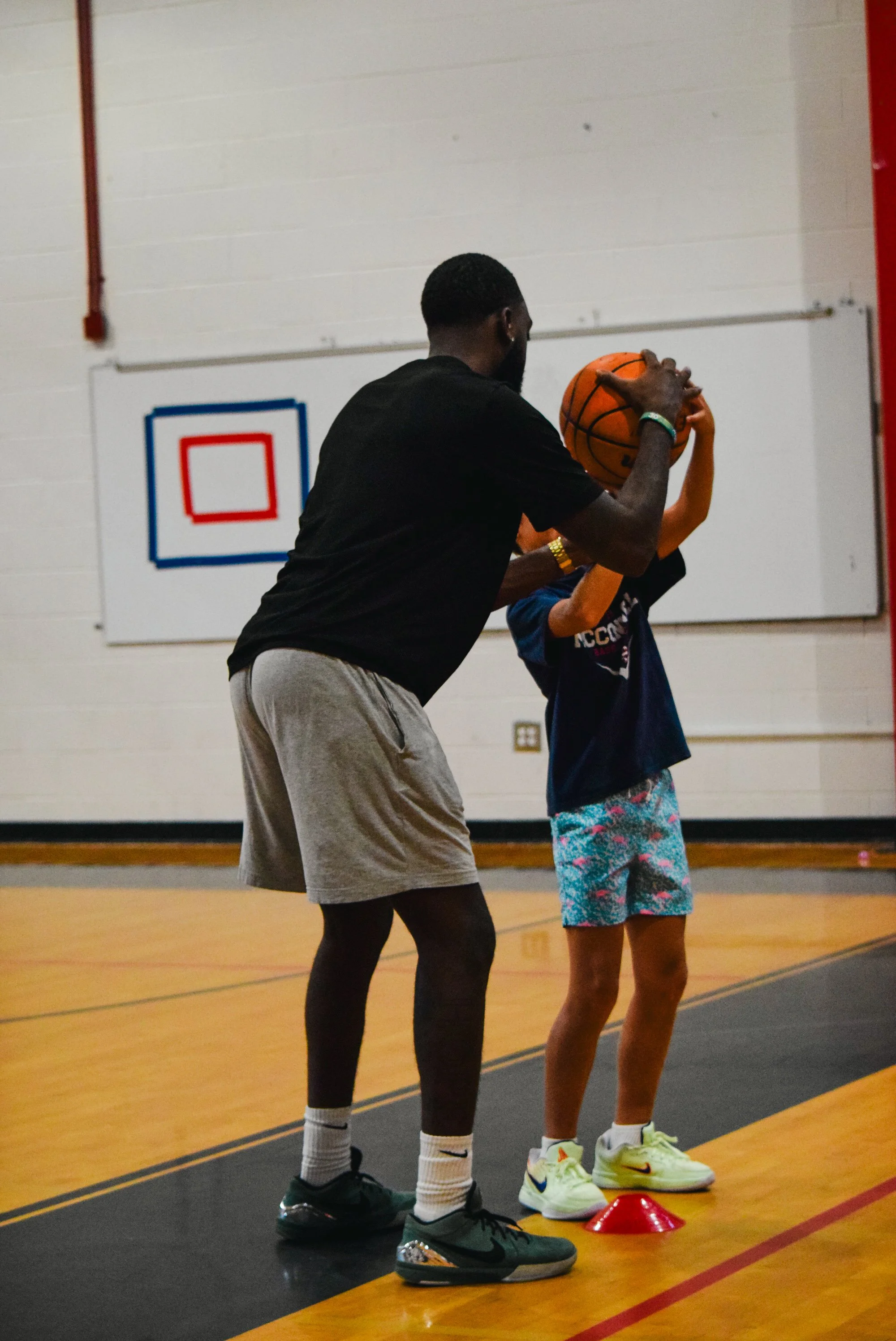 A basketball coach helping a young girl practice shooting on an indoor basketball court.