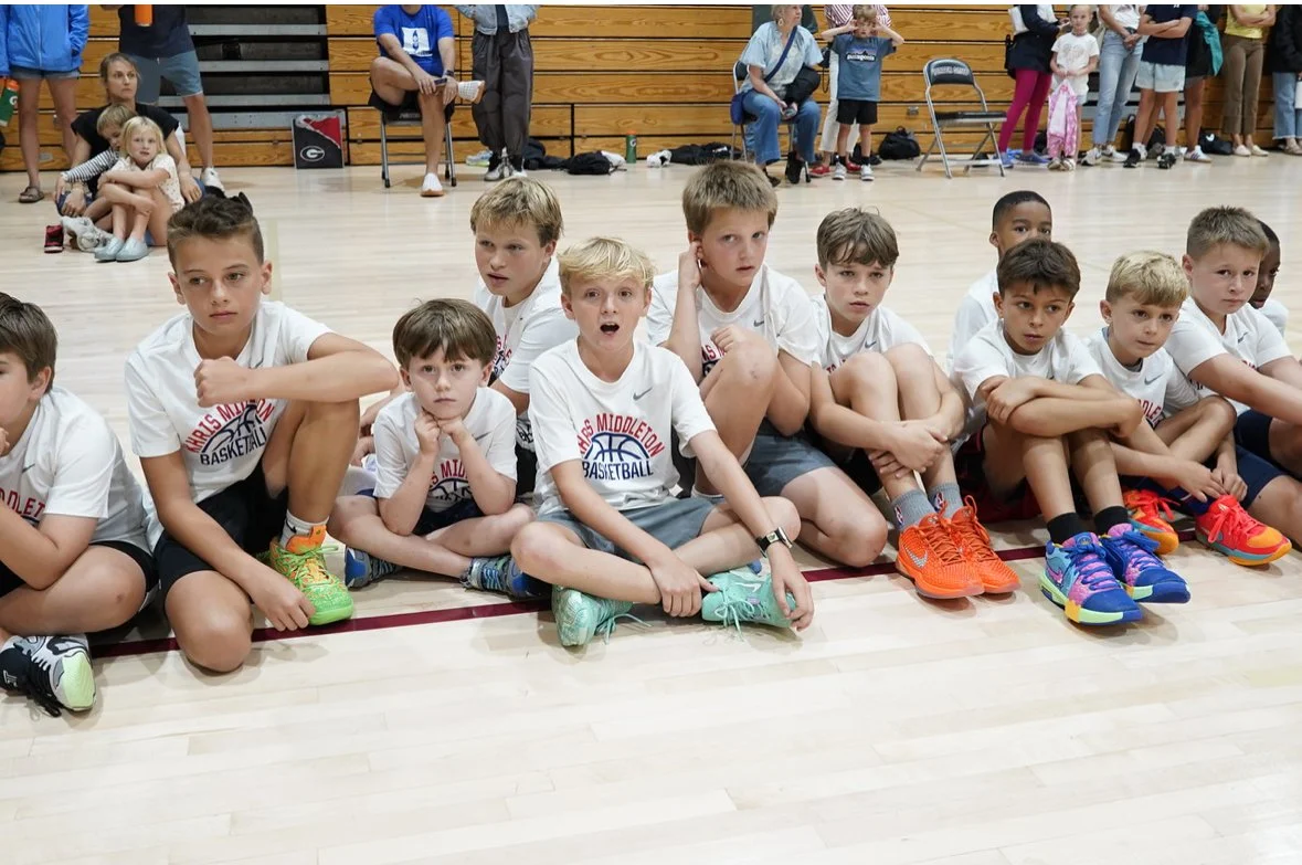 Young boys in white basketball jerseys sitting on gym floor, watching a basketball game, with spectators standing and sitting in the background.