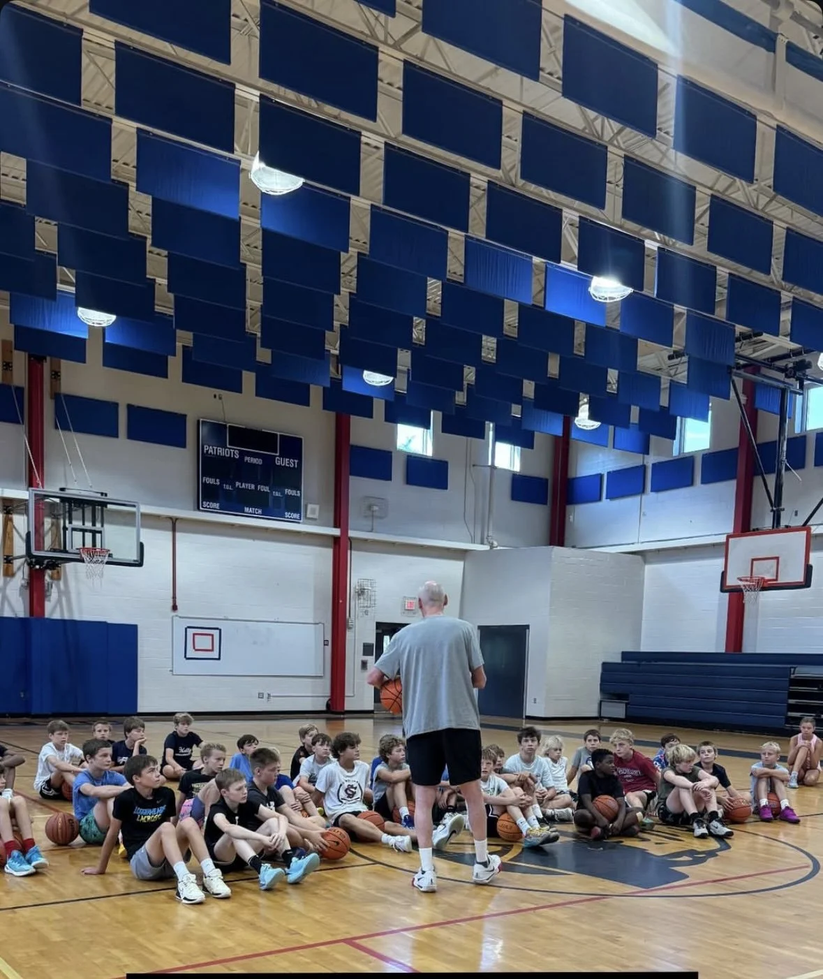 A coach addressing a group of young basketball players sitting on the gym floor inside a gymnasium with basketball hoops and scoreboards.