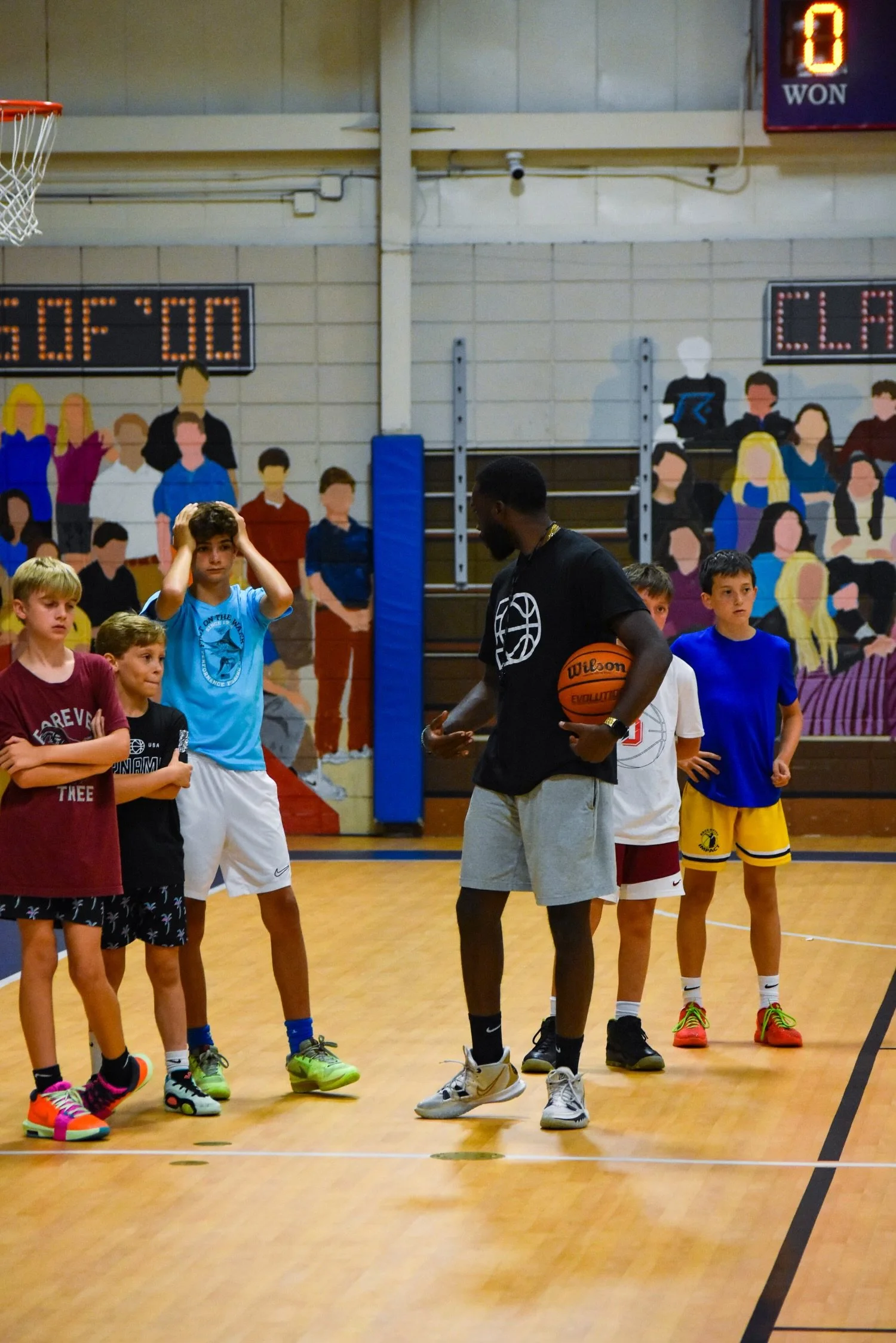 A basketball coach speaks to a group of young boys on an indoor basketball court. The coach holds a basketball, and the boys stand attentively, wearing athletic clothes. Behind them, a colorful mural of a crowd decorates the gym wall, and a scoreboar