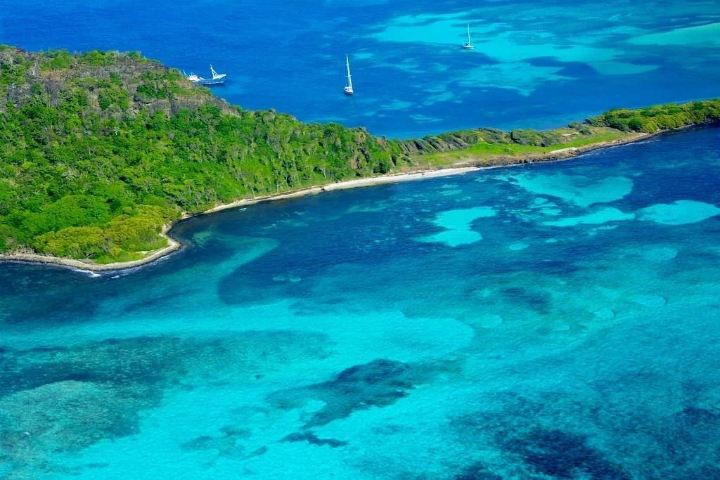 Aerial view of a tropical coastline with a green island, clear turquoise water, and sailboats in the distance.