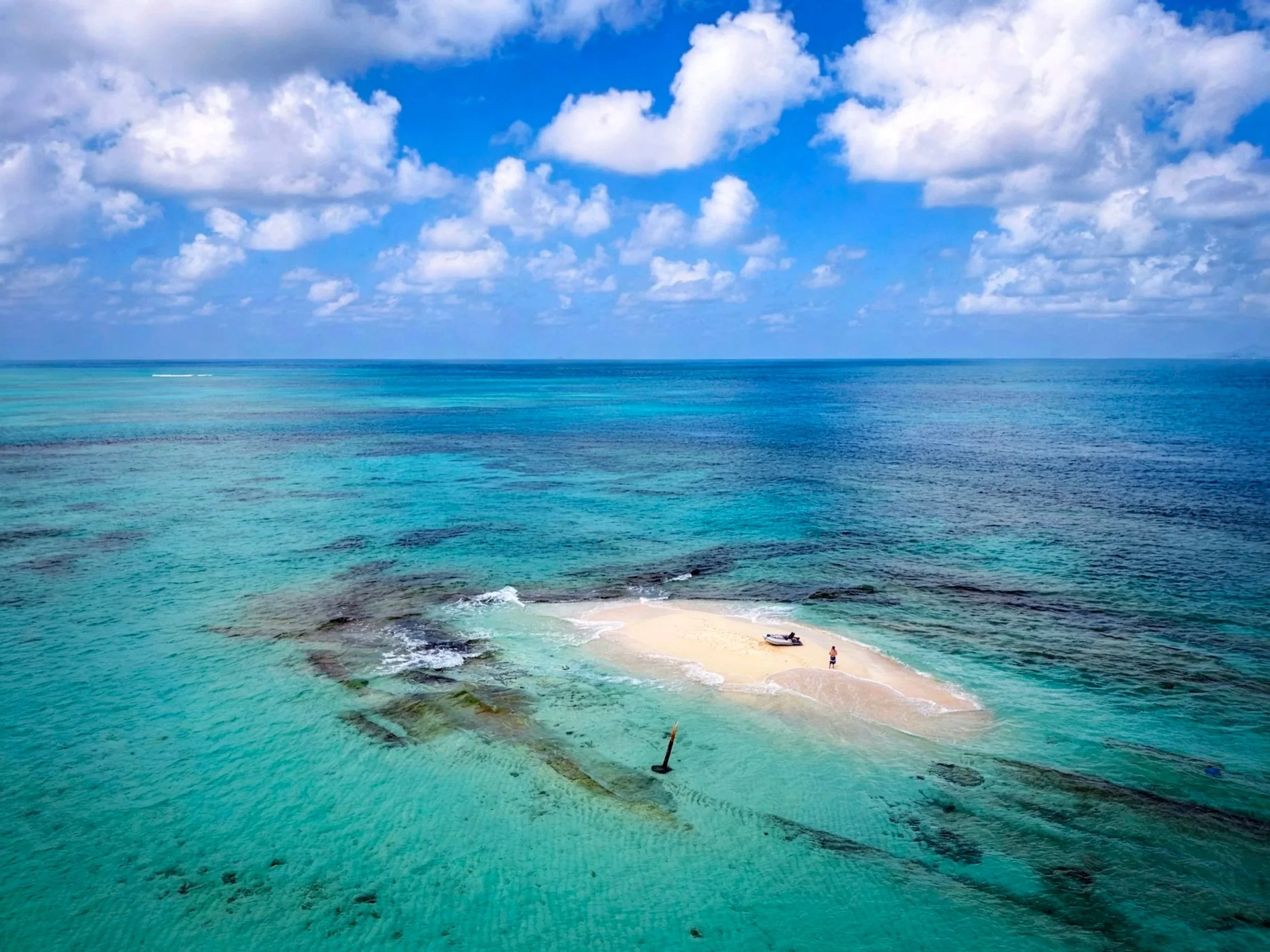 A small sandbar with a person and a boat surrounded by clear turquoise ocean water under a partly cloudy blue sky.