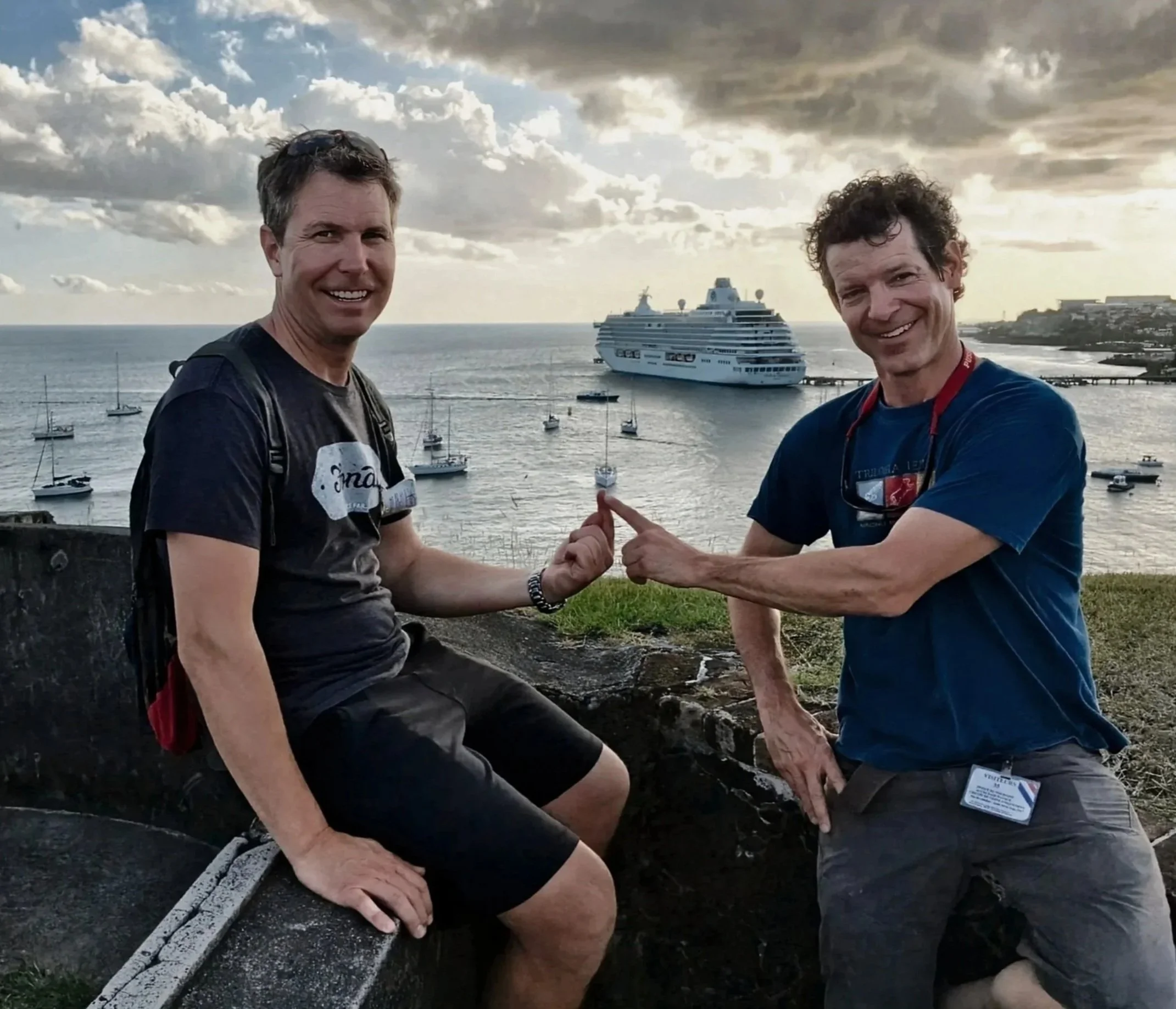 Saltline Sailing captains and founders Matt Hillhouse and Drew Bell smiling and pointing at each other on the water at sunset, with small boats and a cruise ship in the background.