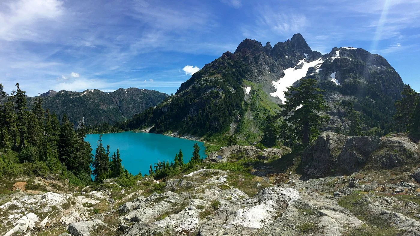 Mountain lake surrounded by pine trees, rocky terrain in foreground, rugged mountain peaks with patches of snow under a partly cloudy sky.