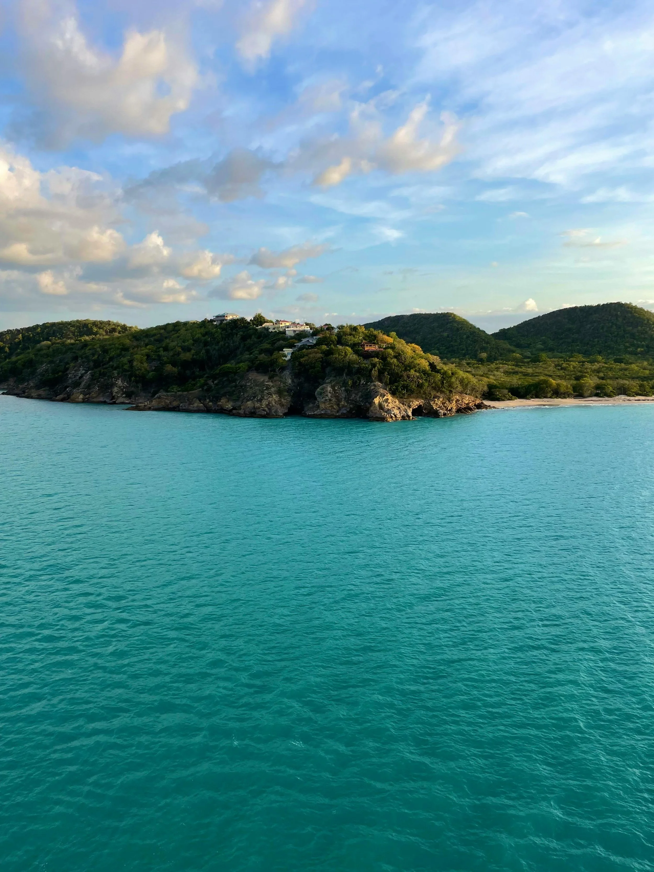 A view of a coastal landscape with turquoise water, rocky shoreline, and green hills with houses on top, under a partly cloudy sky.