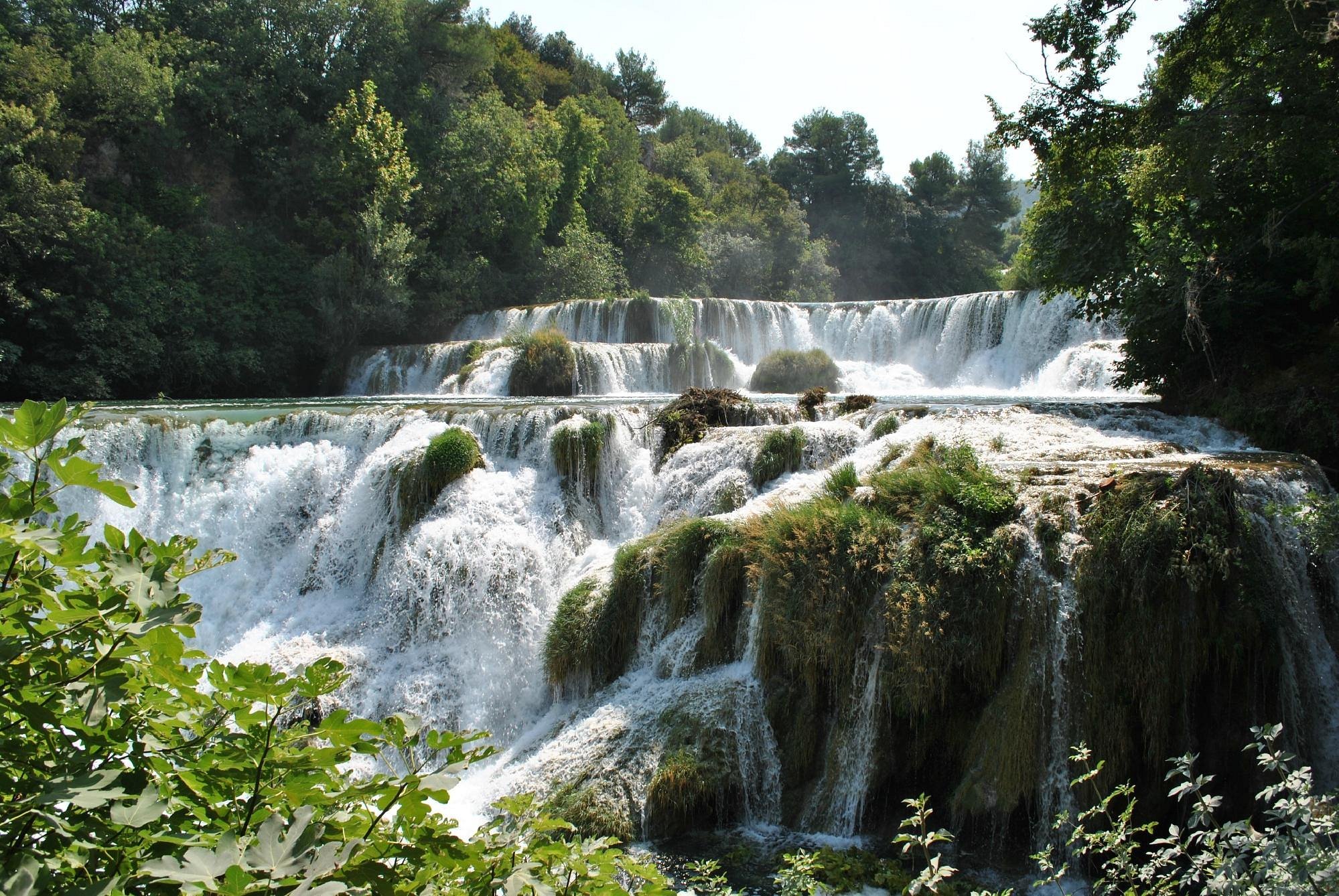 A multi-tier waterfall flowing over rocks, surrounded by lush green trees and foliage on a sunny day.