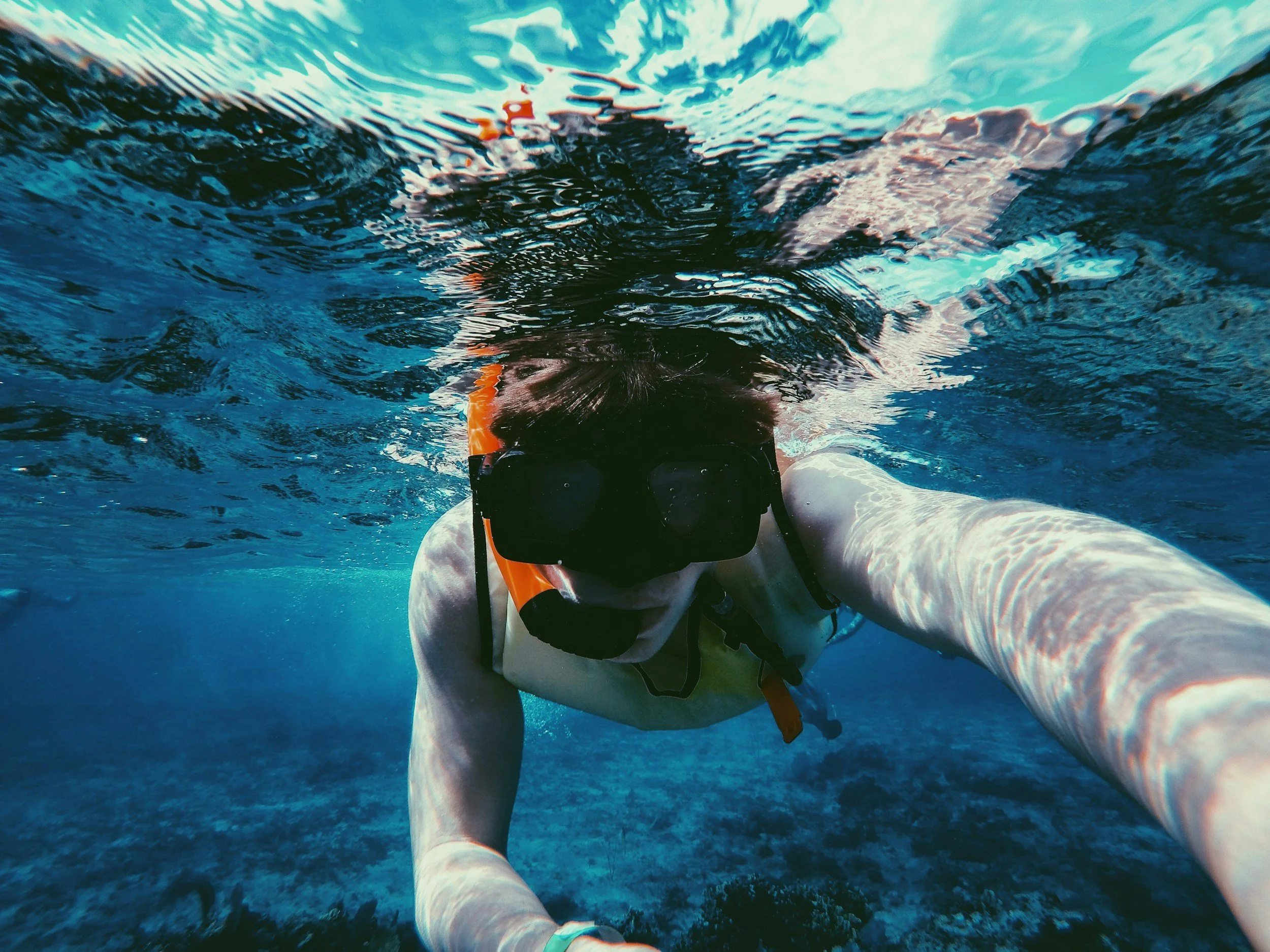 A person snorkeling underwater, wearing a black mask and snorkel, with their arm extended towards the camera, in a clear blue ocean.