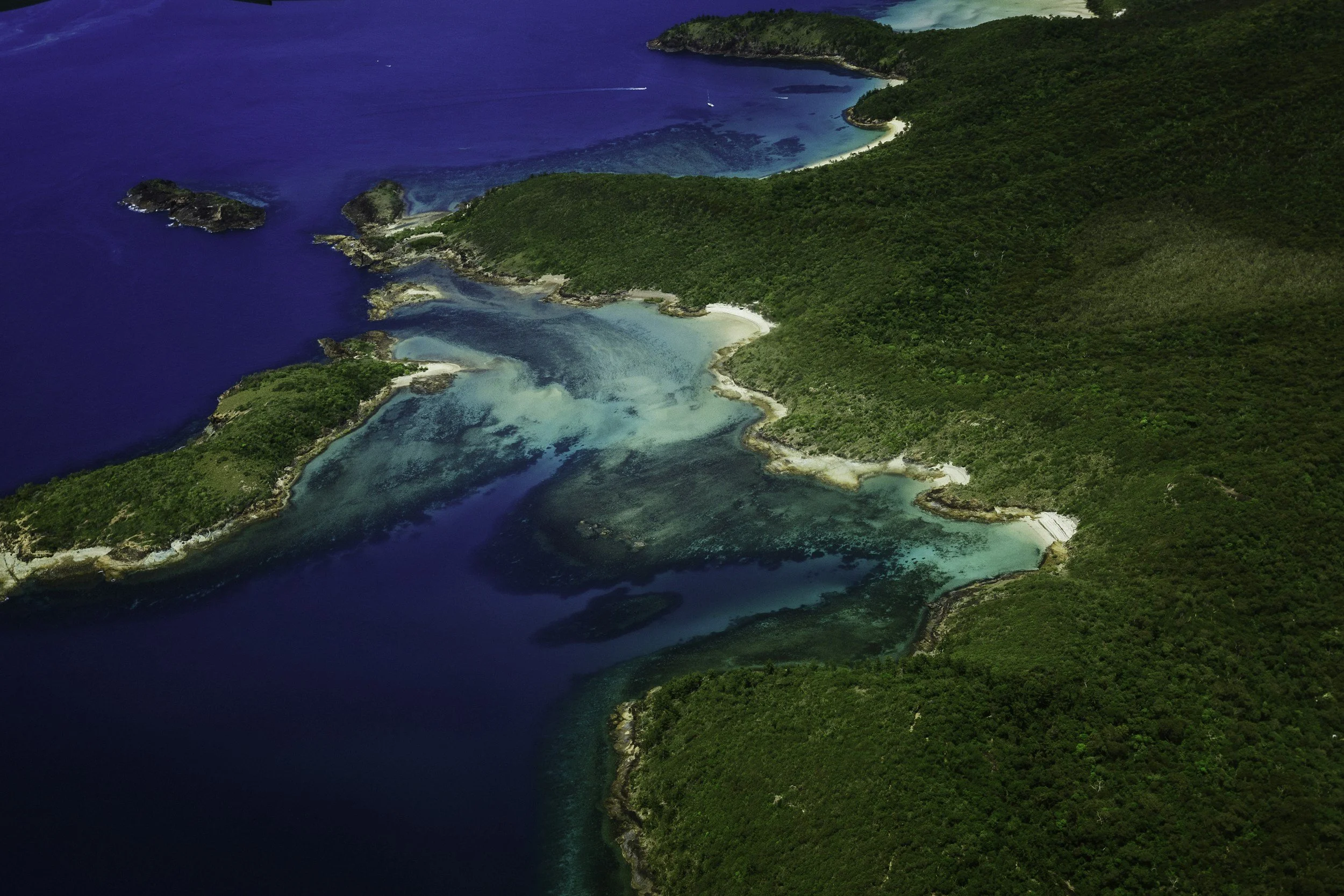 Aerial view of a tropical coastline with lush green forests, white sandy beaches, and clear blue water with shallow coral reefs.