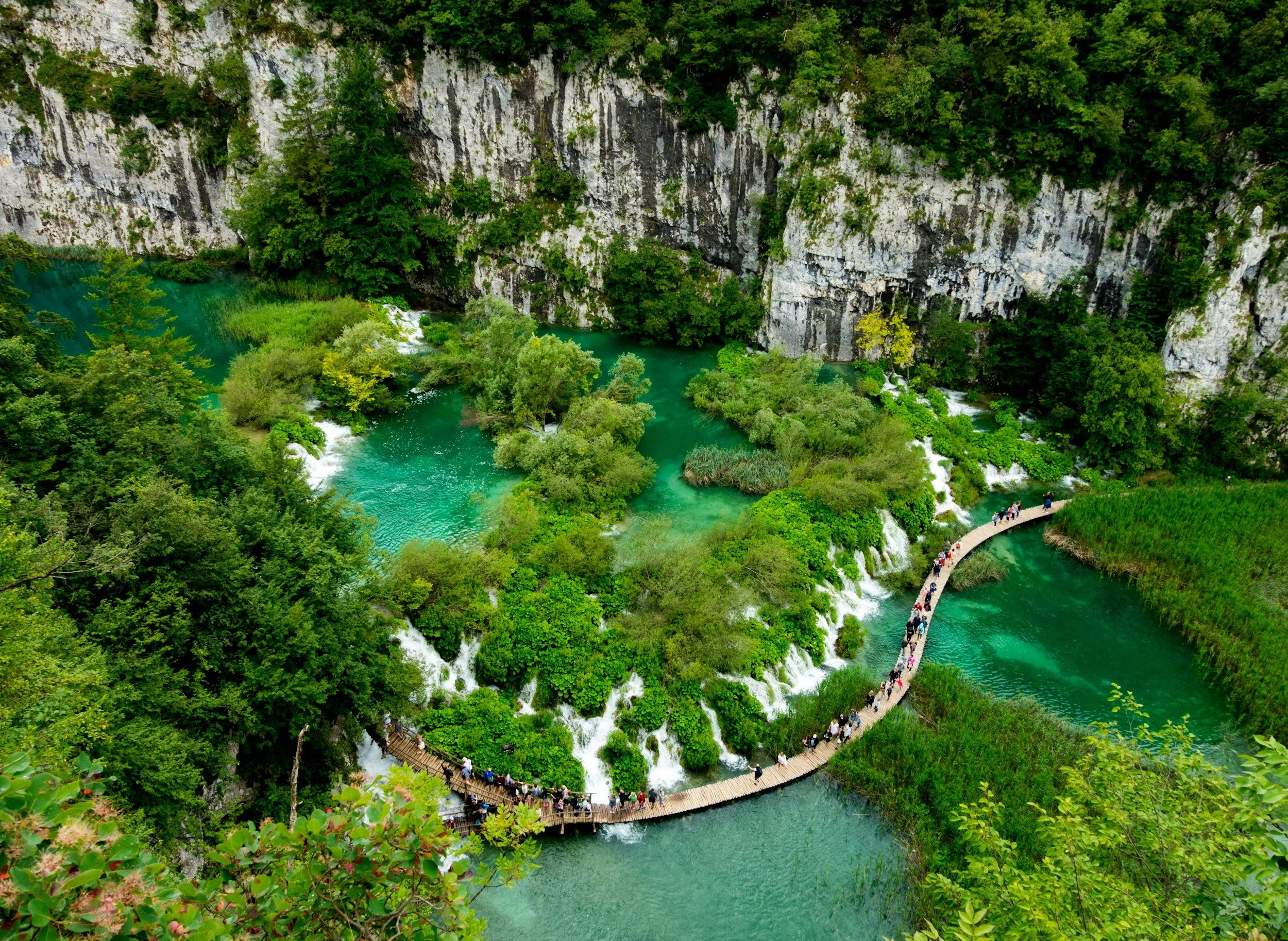 Aerial view of a lush green landscape with waterfalls, turquoise lakes, and a winding wooden footbridge with many people walking on it in Split, Croatic.