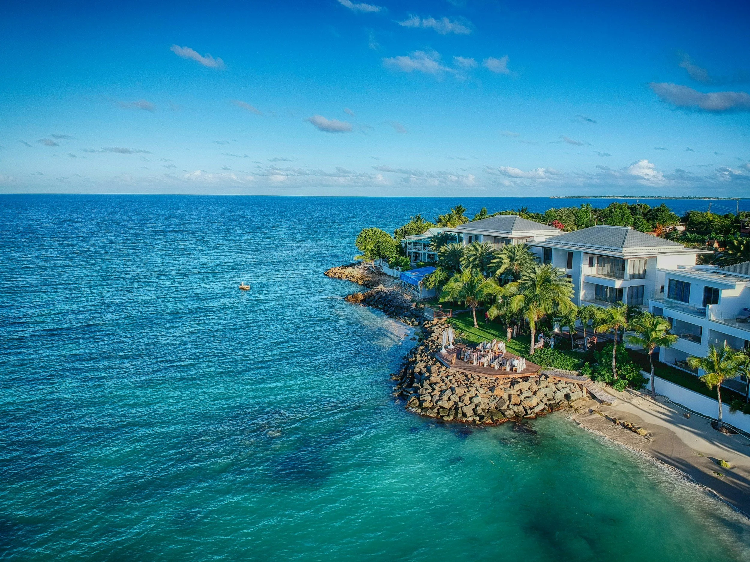 Aerial view of a beachfront resort in Antigua and Barbuda with modern white buildings, palm trees, and oceanfront dining on a wooden deck above a rocky shoreline, overlooking clear turquoise Caribbean water under a partly cloudy sky.