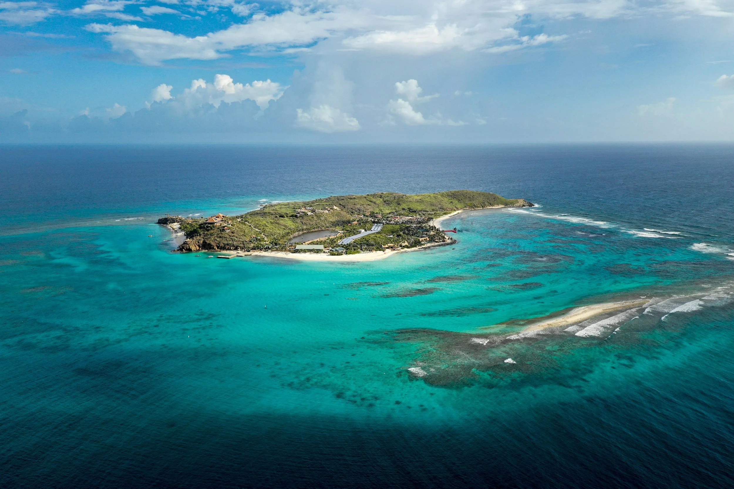 Aerial view of a British Virgin Islands tropical island with a white sand shoreline, lush green vegetation, and turquoise Caribbean water under a partly cloudy sky, perfect for liveaboard sailing routes and ASA certification voyages.