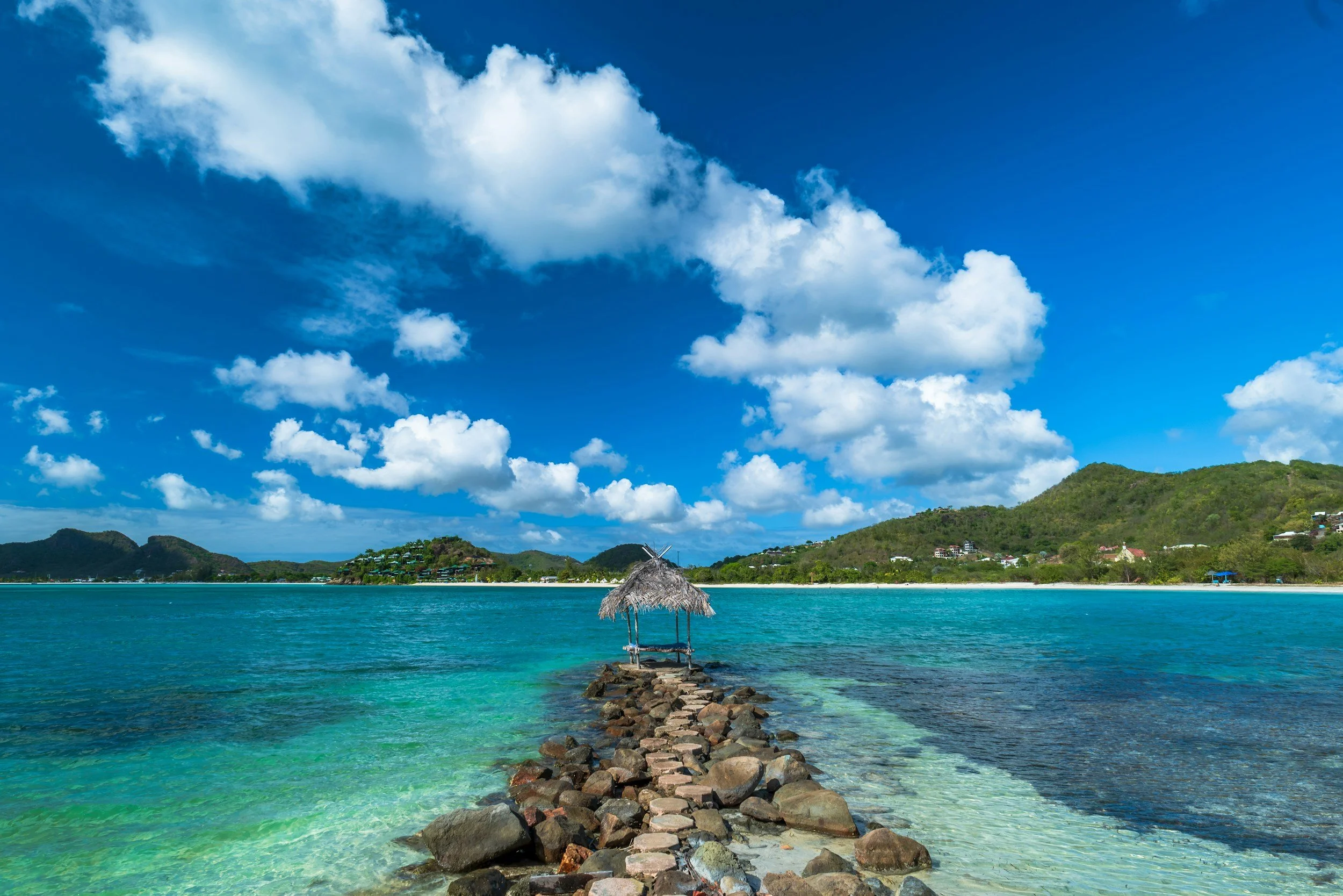 Tropical beach in Antigua and Barbuda with a stone pathway leading to a thatched overwater hut, turquoise shoreline, lush green hills, and a bright blue sky with scattered white clouds.