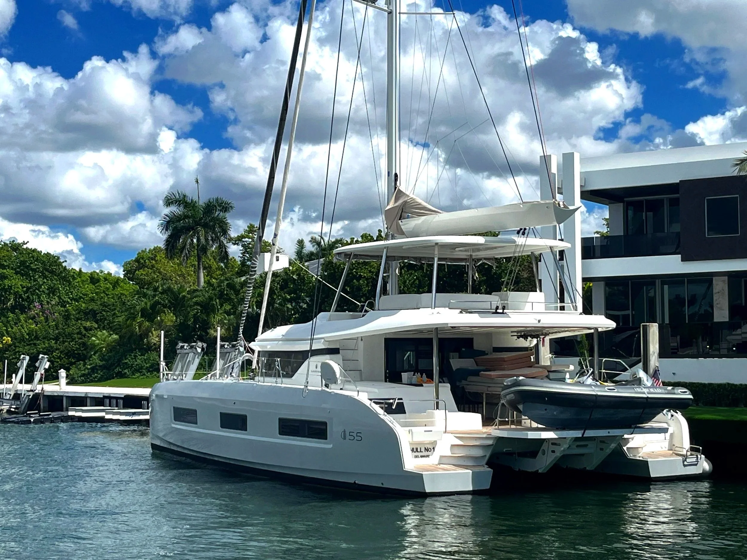 Lagoon 55 luxury catamaran docked at a marina with a modern waterfront home and partly cloudy sky, ideal for ASA certified liveaboard sailing training and adventure charters.