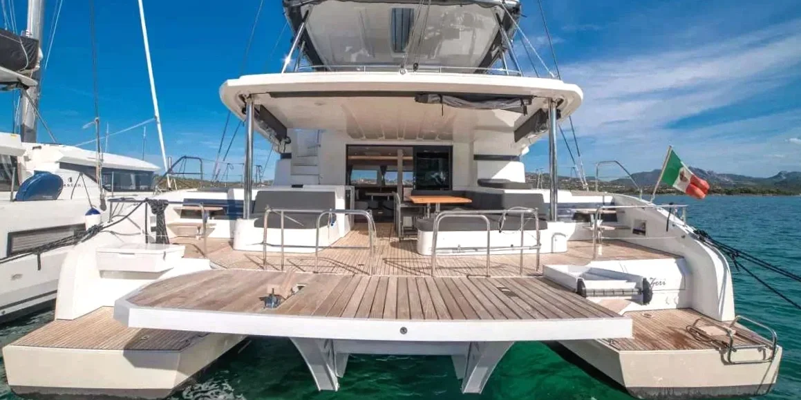 Deck of a luxury yacht with outdoor seating area, Italian flag at the stern, and clear blue sky and water in the background.