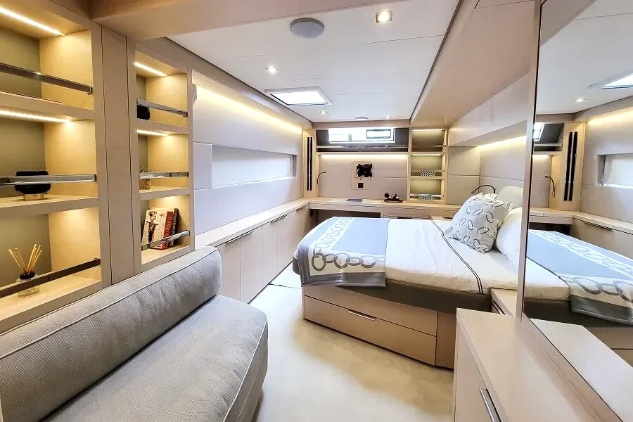A modern bedroom inside the Lagoon 55 luxury catamaran yacht with beige walls and built-in shelving, a bed with white and beige linens, a sofa, and mirrored closet doors.