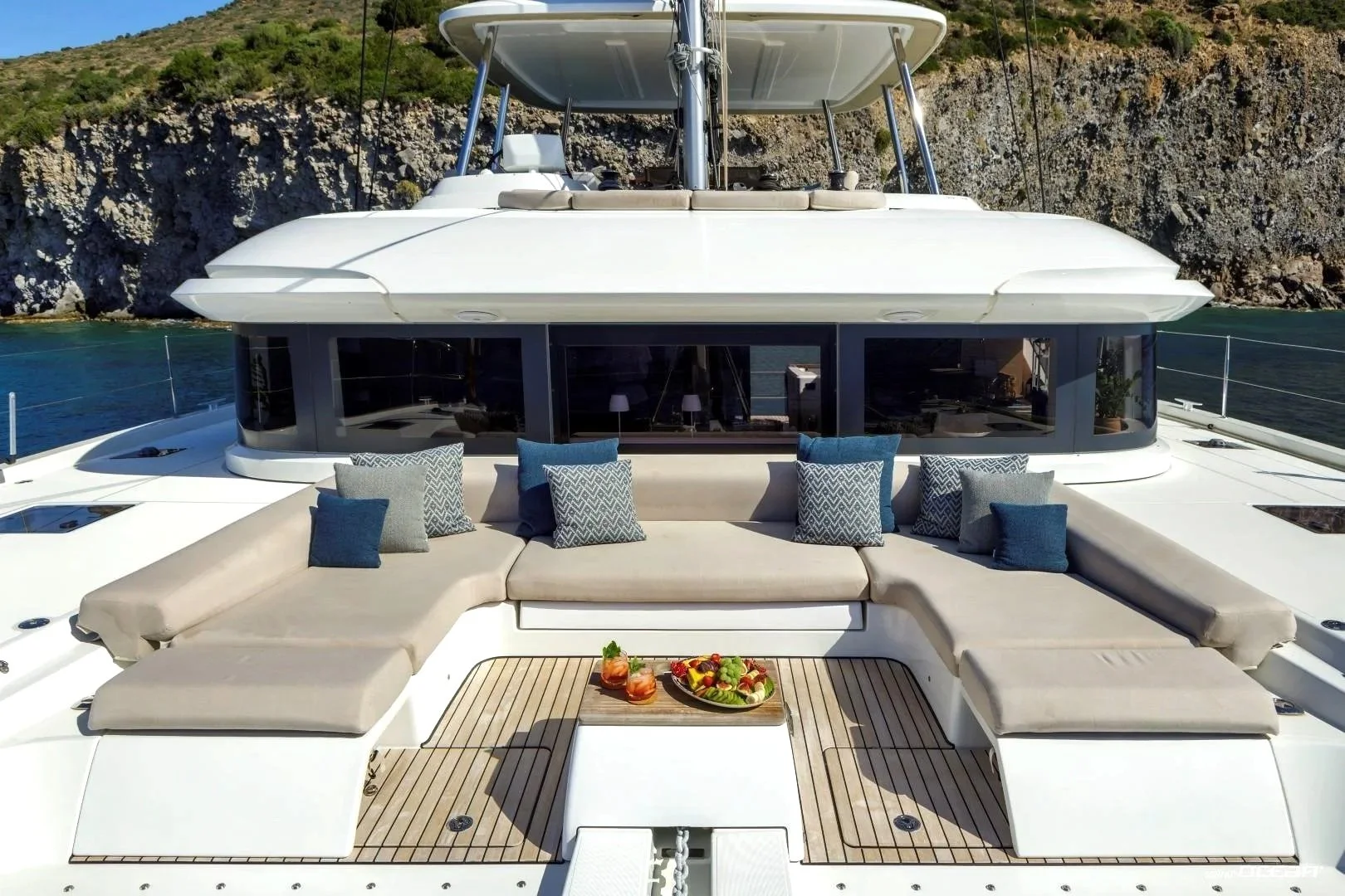A luxury yacht with beige cushioned seating, decorated with blue and patterned throw pillows, positioned on a wooden deck with a fruit platter and drinks, with rocky hills and water in the background.