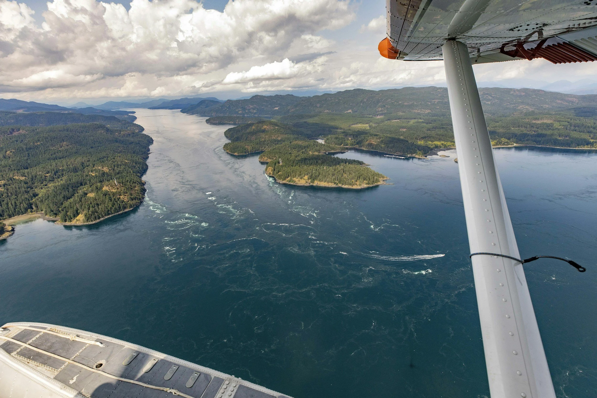 Aerial view from a small airplane showing the Pacific Ocean cutting through the forested landscape of British Columbia with islands and hills under a partly cloudy sky.