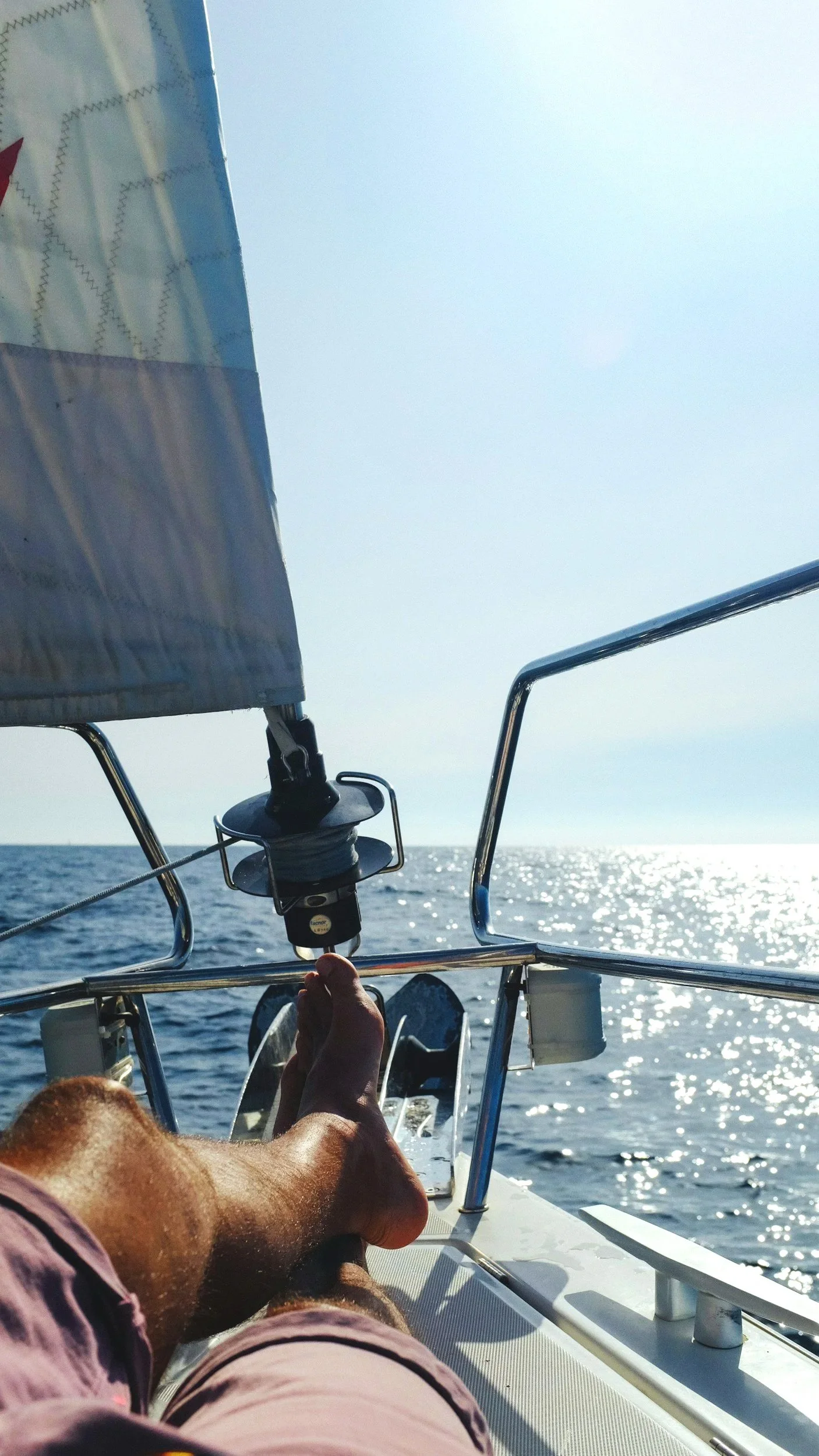 A person relaxing on a sailboat during daytime, with one leg stretched out and the ocean in the background, sunlight reflecting on the water.