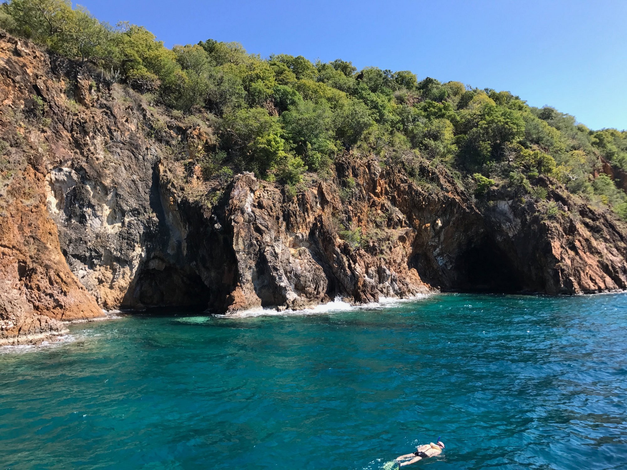 A person snorkeling in a clear blue body of water near a rocky shoreline with a steep, green, forested hillside.