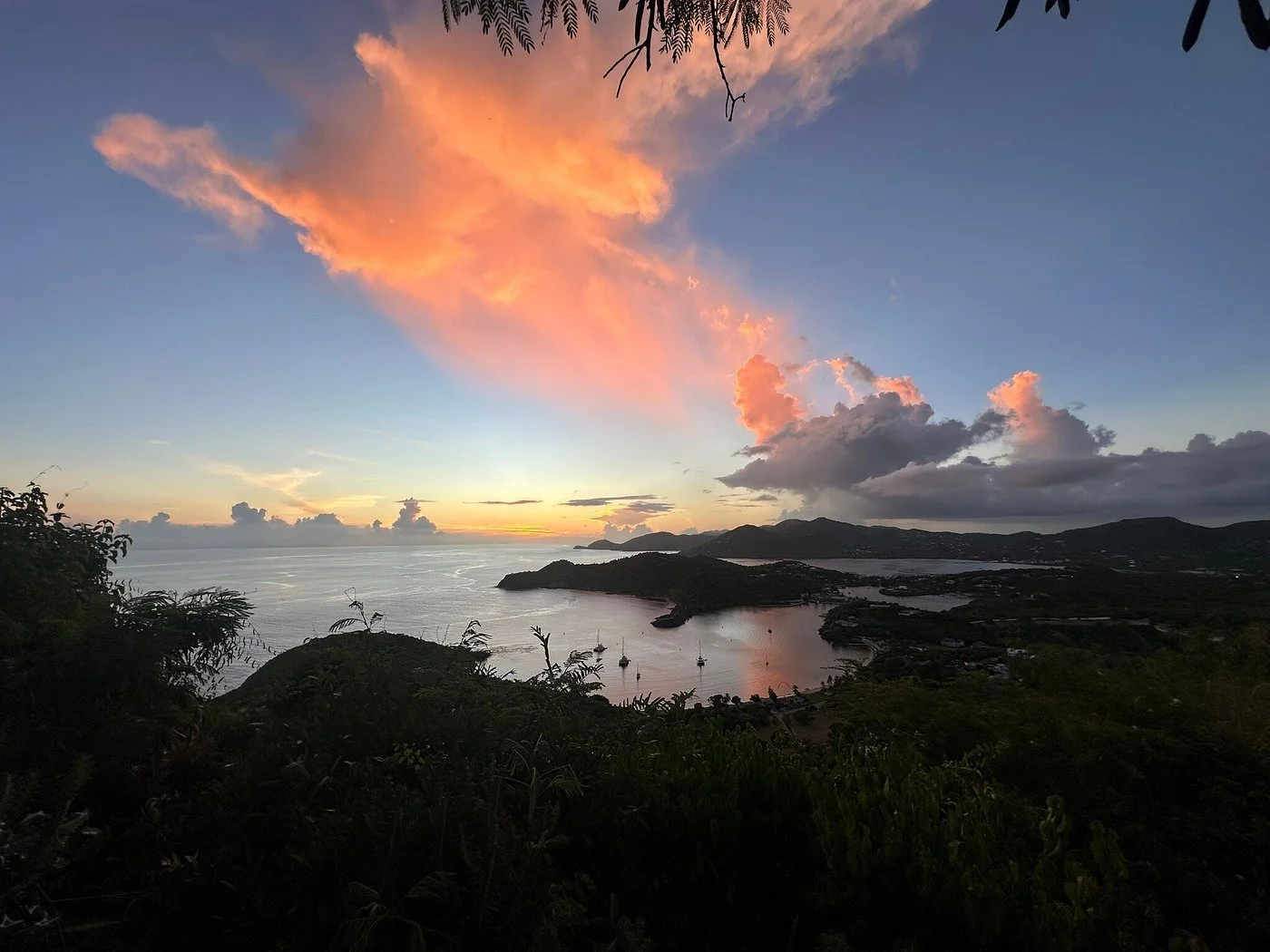Sunset over a bay with boats, islands, and mountains in the distance, with pink and orange clouds in the sky.