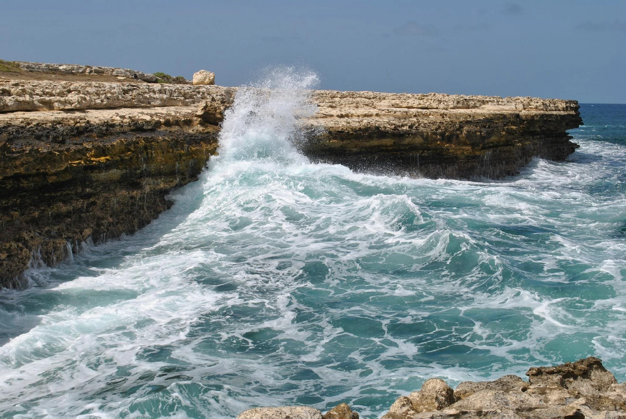 Ocean waves crashing against rocky shore with clear blue sky.