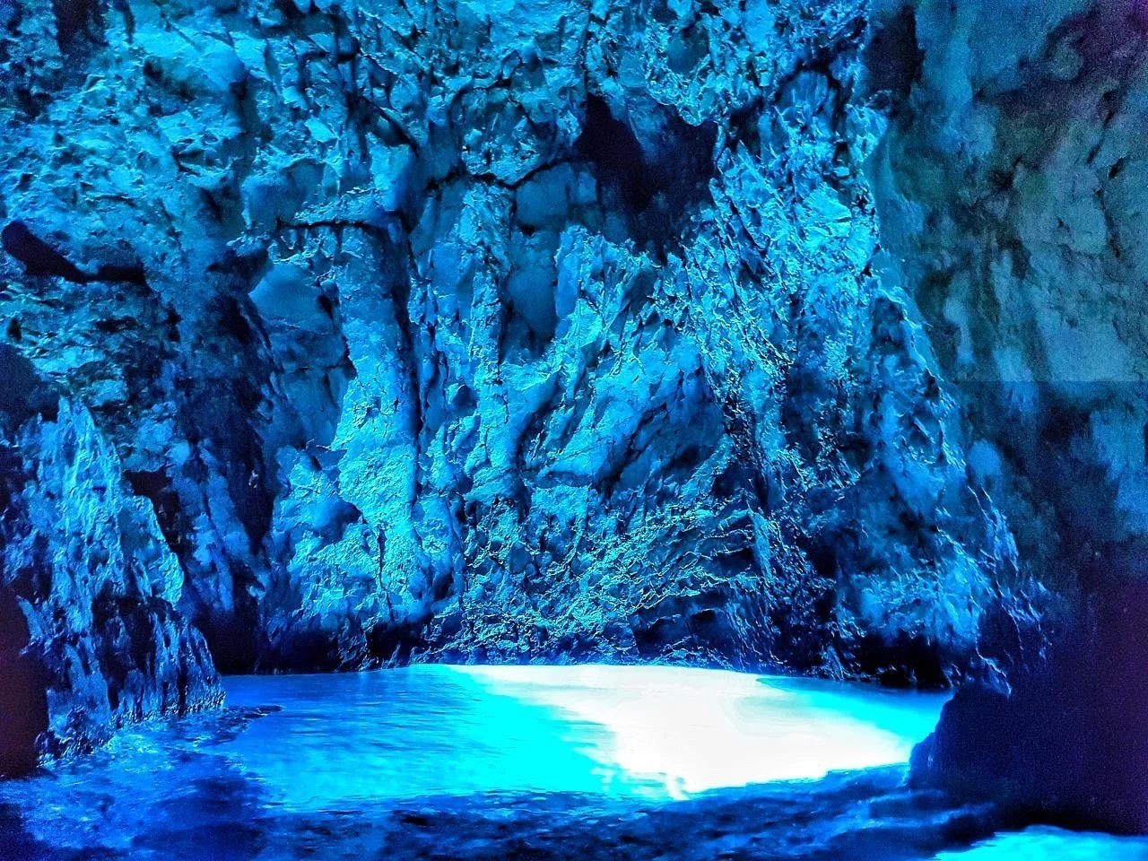 Blue-lit ice cave with rocky walls and a partially frozen body of water inside.
