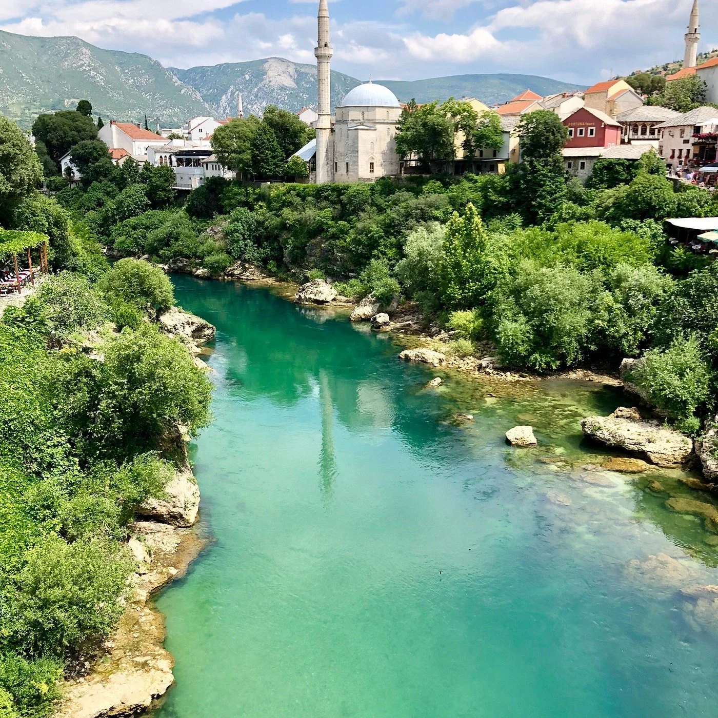 A scenic view of a turquoise river flowing through lush green vegetation with a historic mosque and colorful buildings on a hillside, and mountains in the background.
