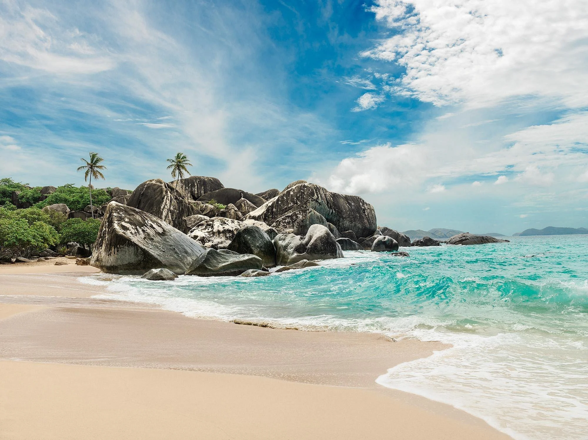 Tropical beach with large rocks, palm trees, turquoise water, and a blue sky with scattered clouds.