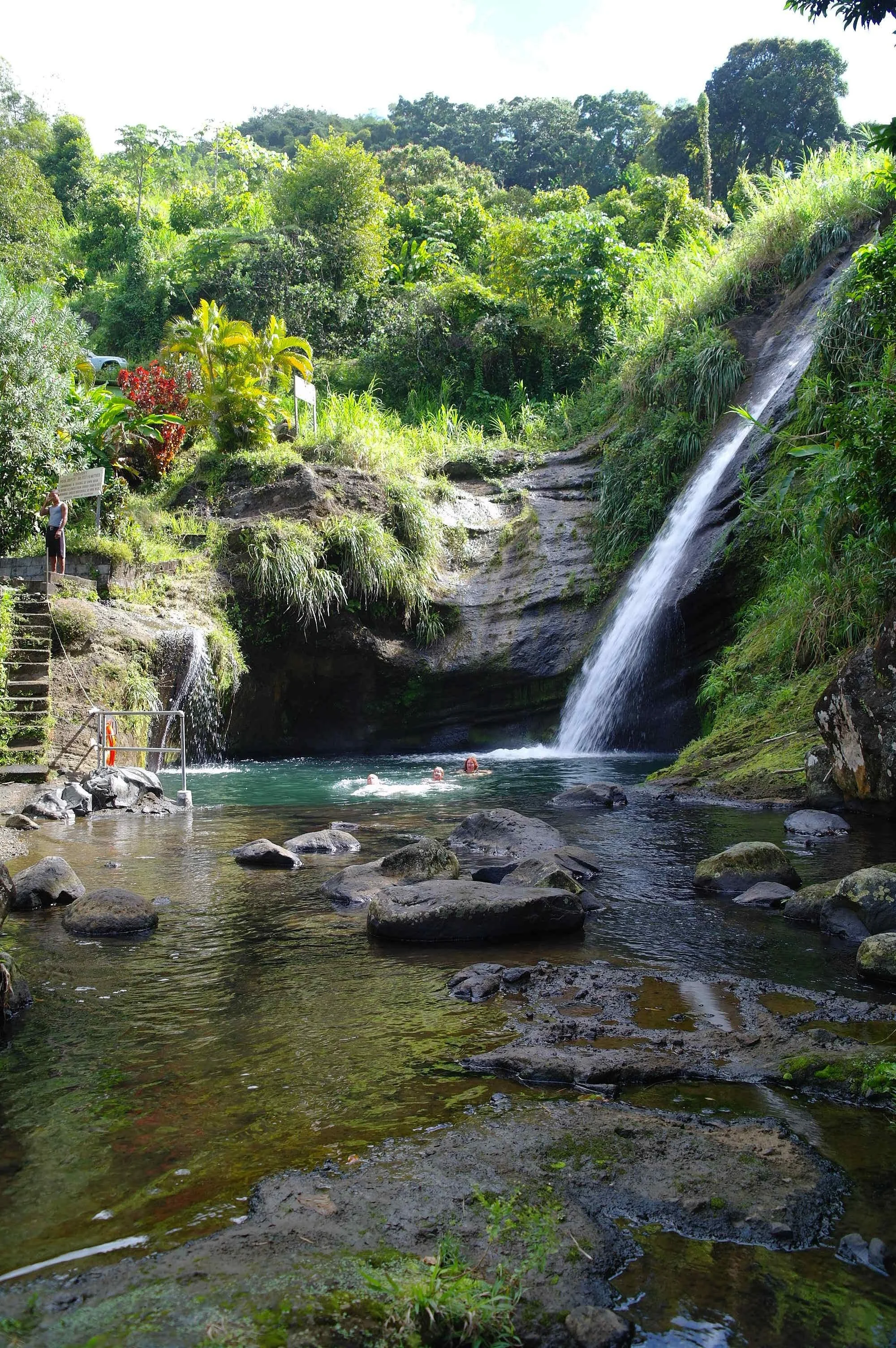 People swimming in a natural pool at a waterfall surrounded by lush greenery and rocks.