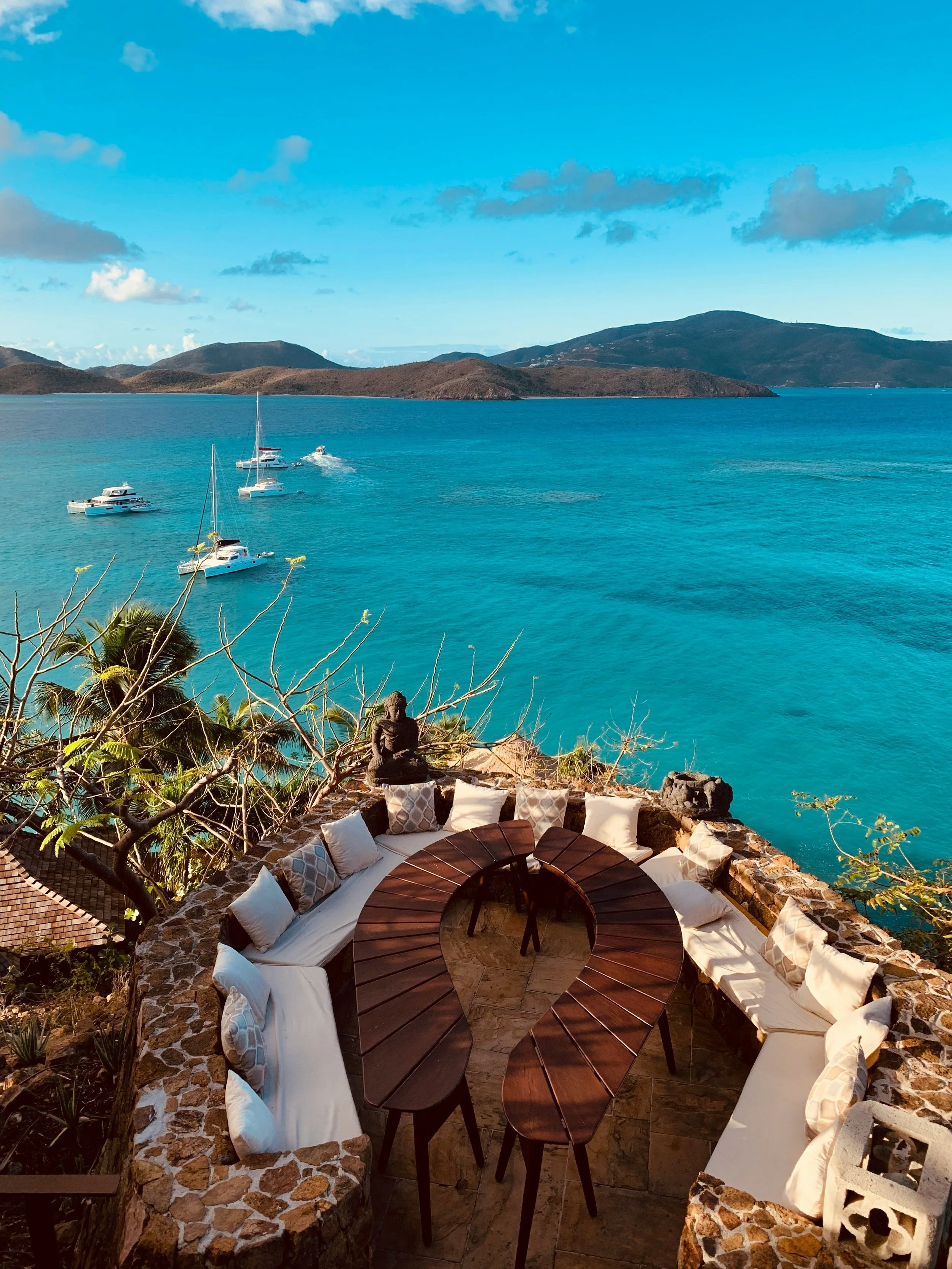 Outdoor seating area with round wooden table and cushioned benches overlooking a turquoise bay with boats, hills, and mountains in the background.
