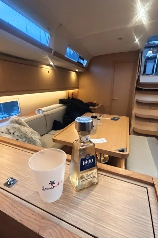 Interior of a yacht or boat with a wooden table, couch with cushions, a door, and stairs, seen through an opening with a water view outside.