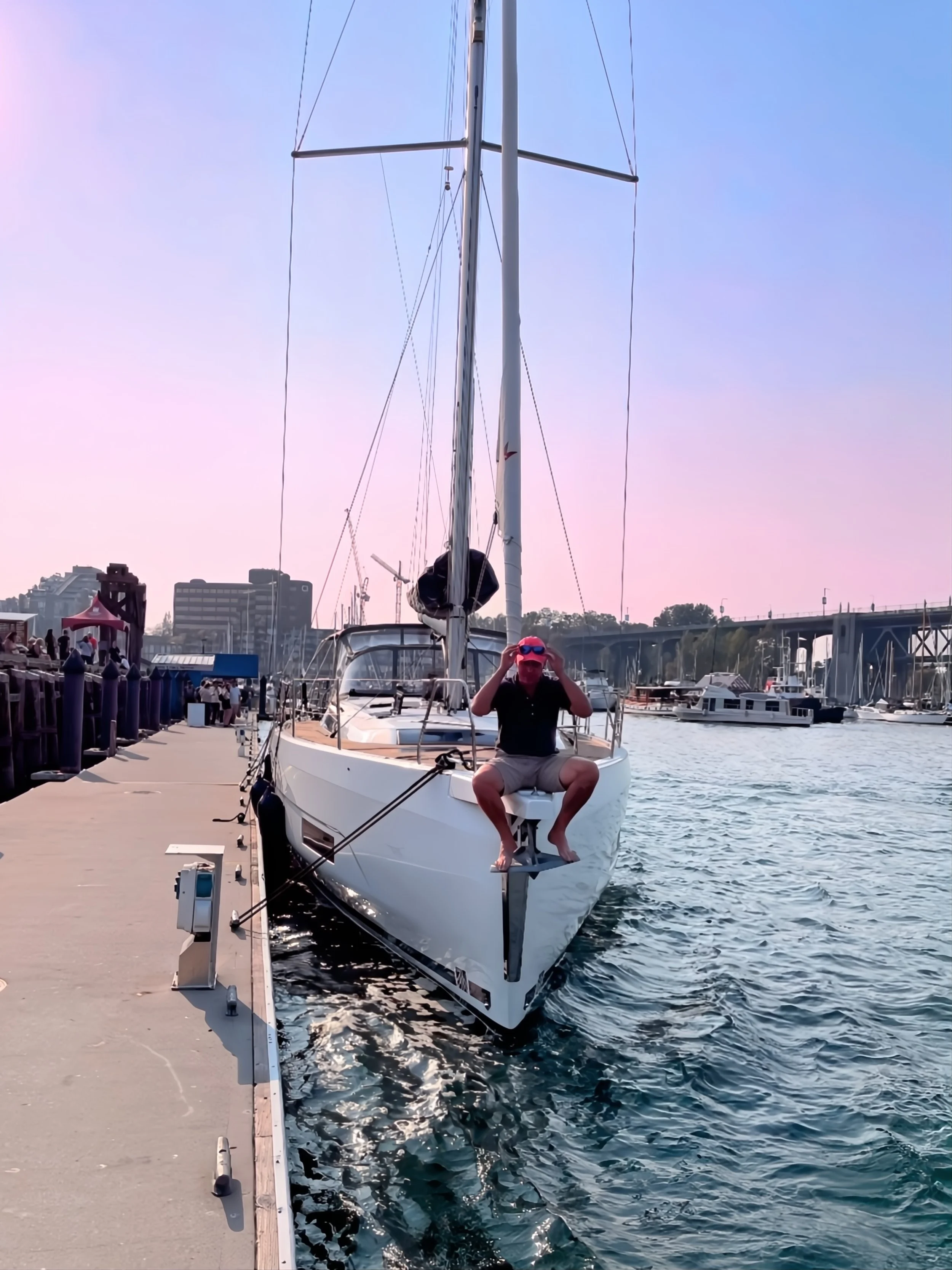 A man sitting on the bow of a white sailboat docked at a marina, adjusting his sunglasses with a pinkish sky in the background.