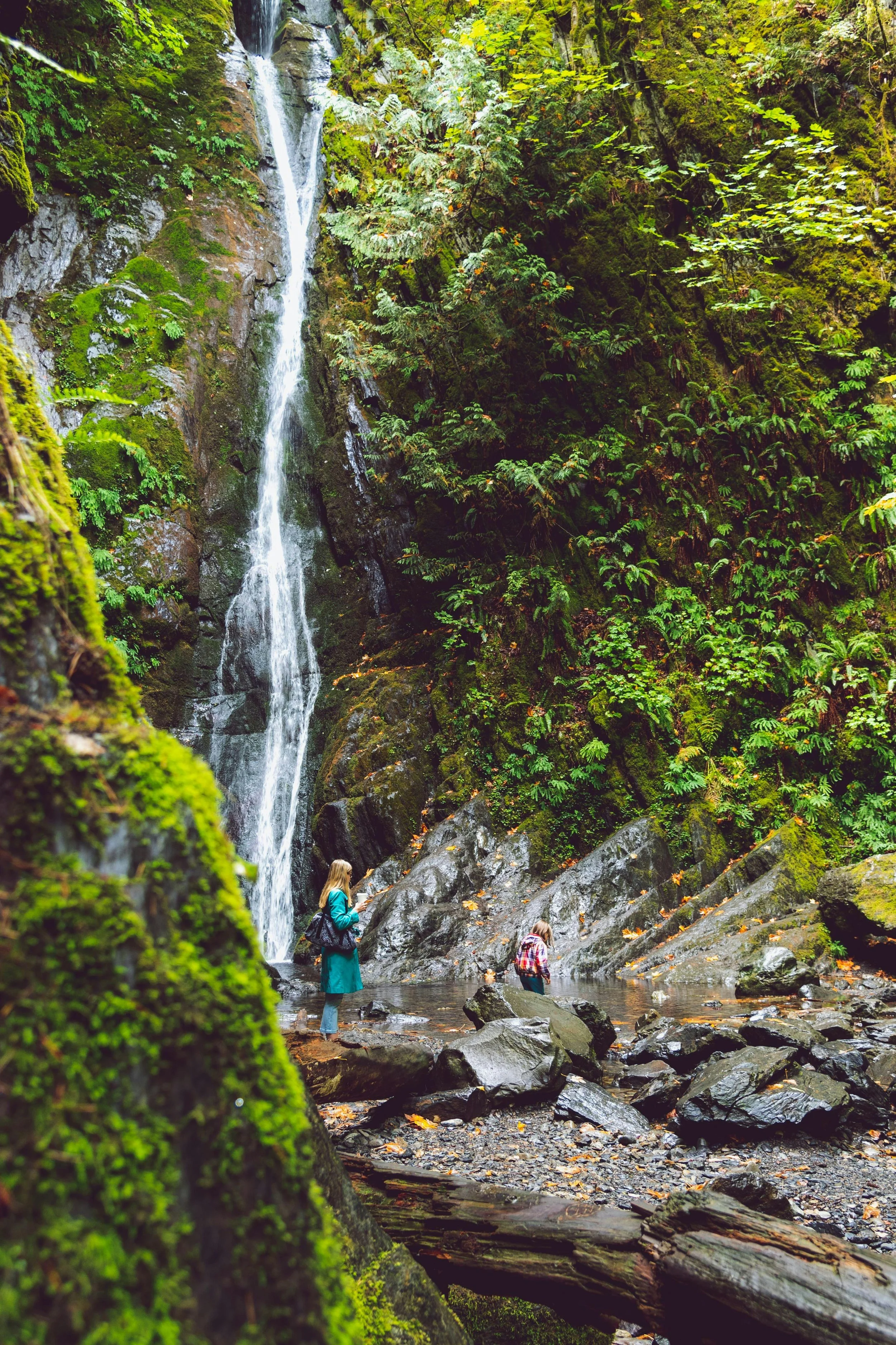 Two hikers standing near a rocky stream at the base of a tall waterfall in a lush, green forest.