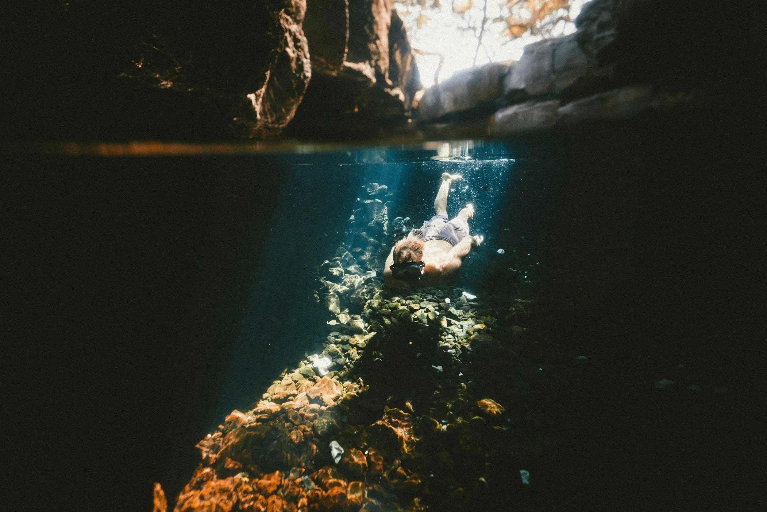 A person swimming underwater in a rocky natural waterway or cave, viewed from below the water surface with rocks at the top and sunlight filtering through.