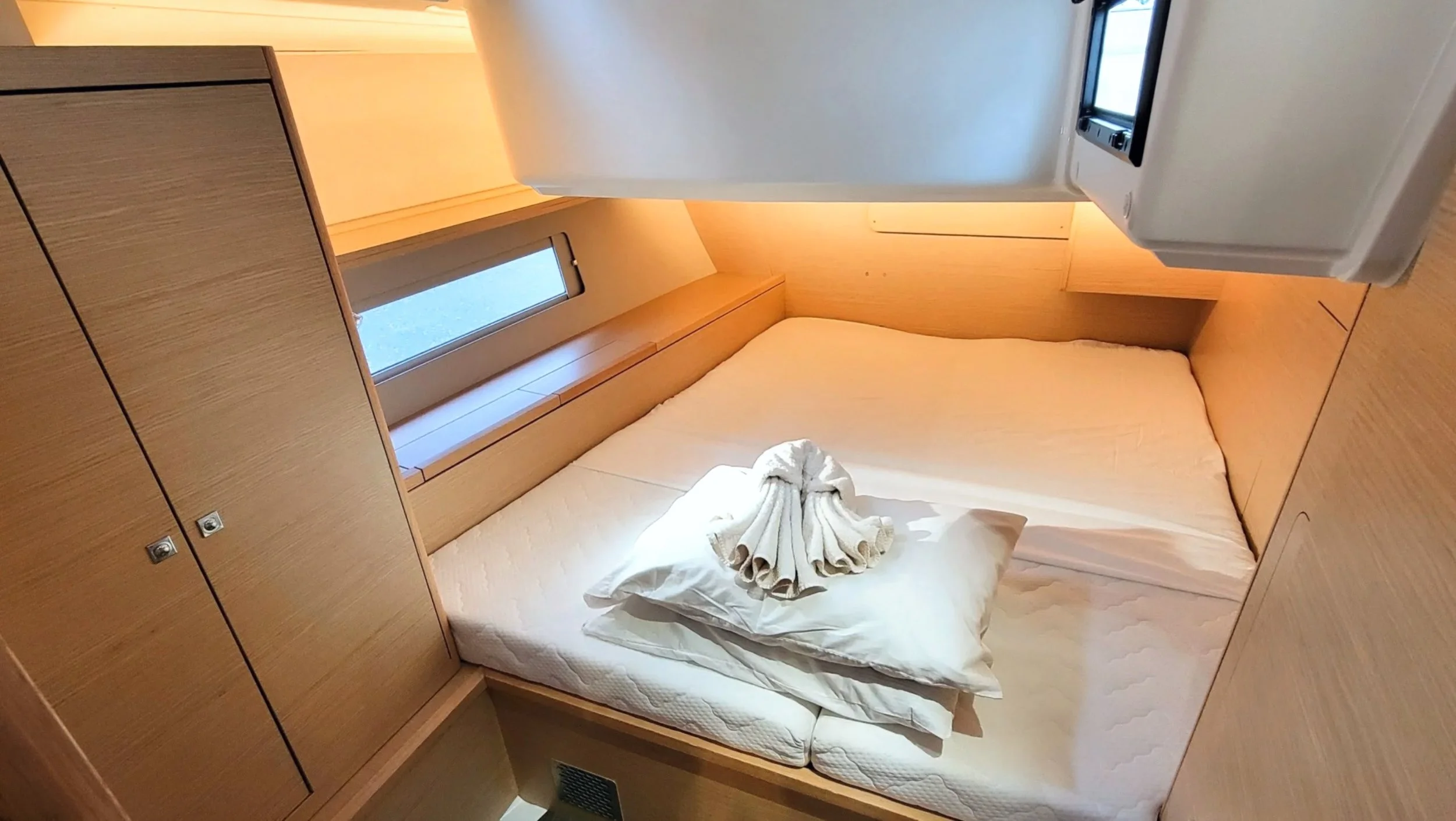 A bedroom with a bed covered in white bedding, a pillow with a towel folded into a swan shape on top, wooden built-in cabinets, a small window, and a television mounted on the wall.