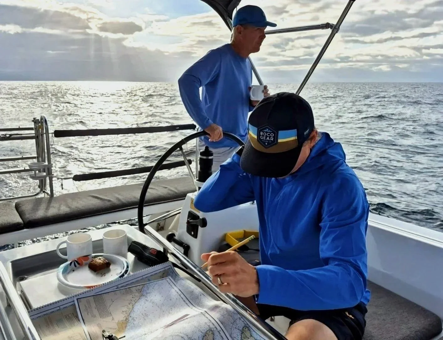 Two men on a boat, one at the steering wheel looking at a map, the other standing in the background holding a mug, over the ocean with a cloudy sky.