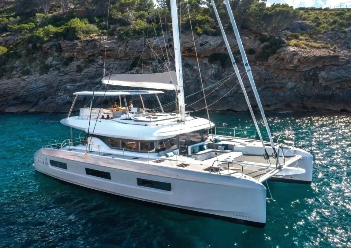 A large white Lagoon 55 catamaran yacht anchored in clear, blue water near rocky shorelines with green vegetation, under bright sunlight.