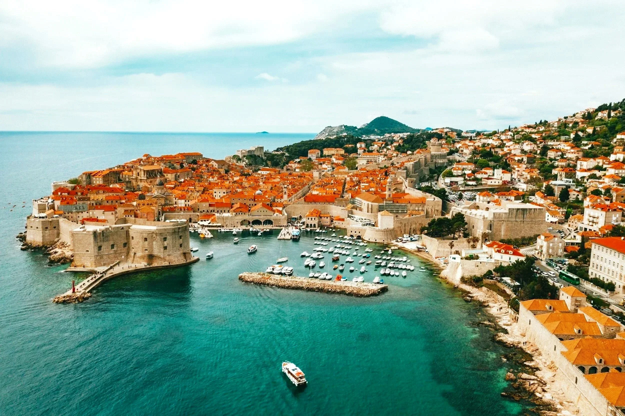 Aerial view of the historic coastal city of Split, Croatia, with red-tiled rooftops, stone walls, and a harbor filled with boats, overlooking a clear blue sea and distant hills.