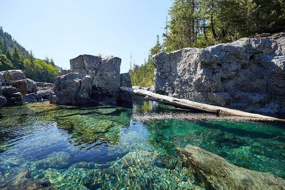 Clear mountain river with large rocks and surrounding forested hills under a bright sky.