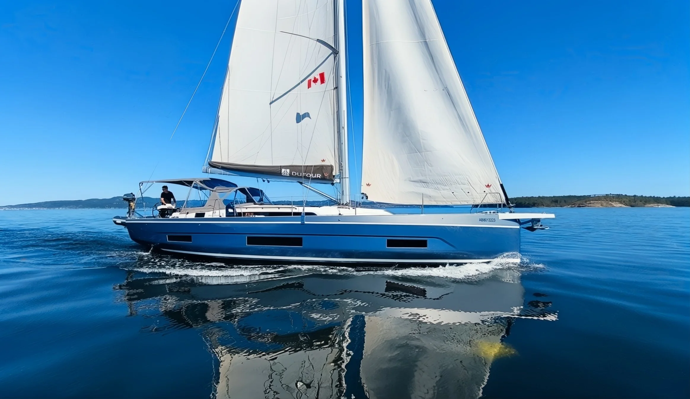 A blue sailing yacht with white sails on calm water near a distant shoreline on a clear, sunny day.