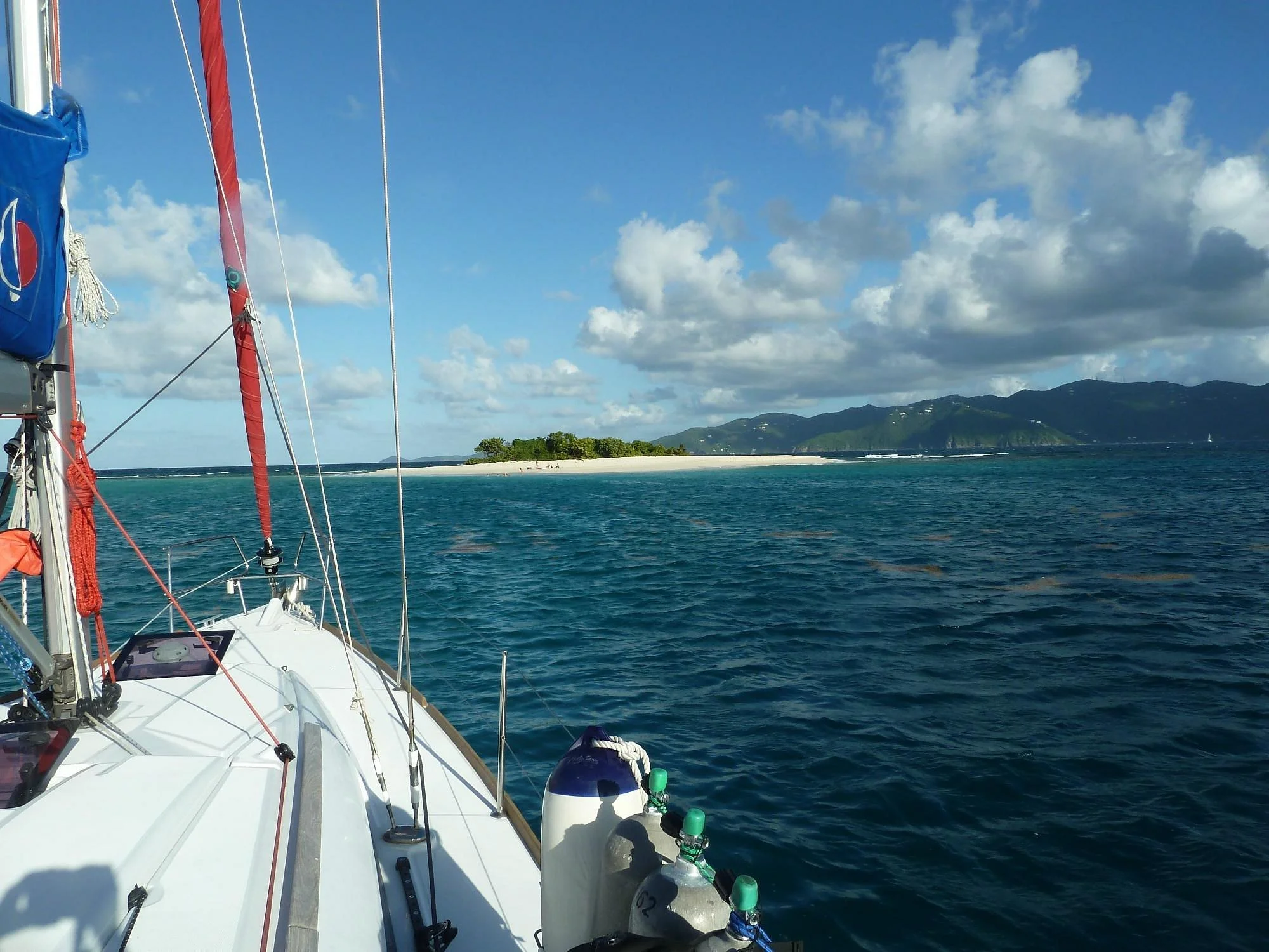 View from a sailboat showing an island with sandy beach, green trees, ocean water, and mountains in the background under partly cloudy sky.