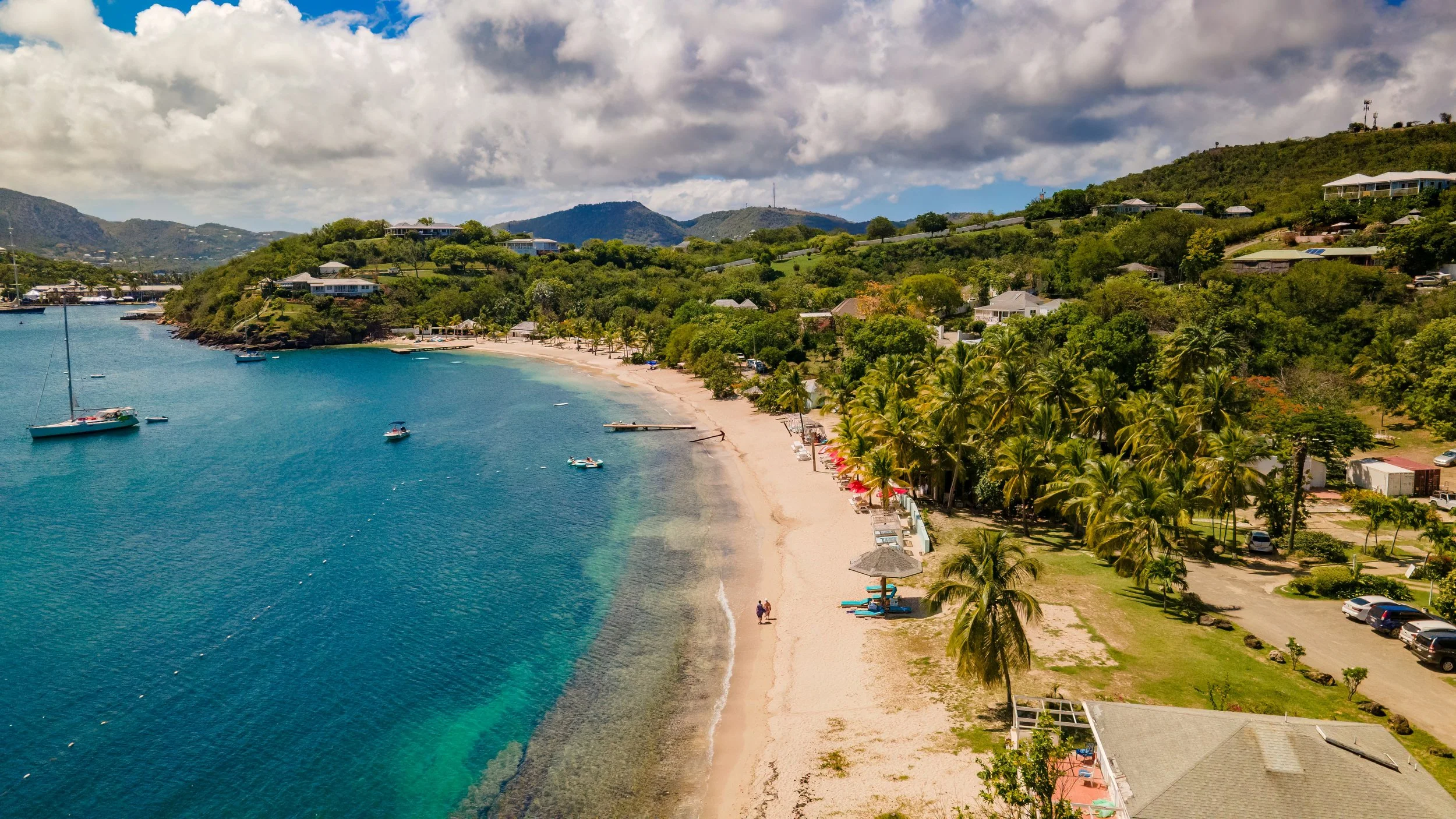 A scenic aerial view of a tropical beach with clear blue water, sailboats, and small boats anchored near the shore. The sandy beach is lined with palm trees, umbrellas, and beach chairs. Hills with lush greenery and houses are visible in the backgrou